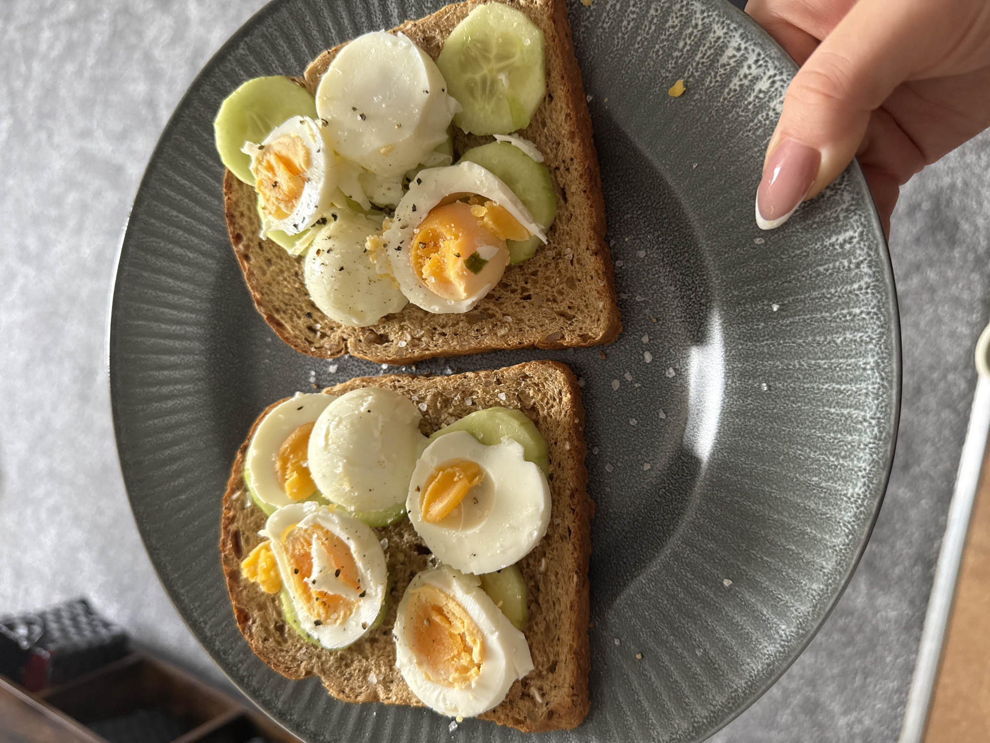 A close-up, high-angle shot captures a pair of egg and cucumber toasts on a dark gray ceramic plate, held by a person's hand. The plate features a textured surface with subtle ridges along the rim. Two slices of whole-grain or whole-wheat bread serve as the base for the toppings. Each toast is generously adorned with thin slices of green cucumber and several pieces of hard-boiled egg. Some eggs are cut in half, revealing their bright orange-yellow yolks, while others are whole circular slices of the white.
The food is seasoned with black pepper, visible as dark specks on the eggs, and coarse salt crystals, which are scattered over the toast, eggs, and the plate itself. The eggs appear to be cooked to a medium-hard consistency, with the yolks still retaining some moisture.
A hand with neatly manicured nails (light pink base with white tips) is seen gripping the right edge of the plate, suggesting the plate is being presented or held up. The background is softly out of focus, showing a light-colored wall or surface on the left and a darker, possibly patterned object or furniture, further back. The lighting is bright and even, indicating a daytime setting. No text is visible in the image.