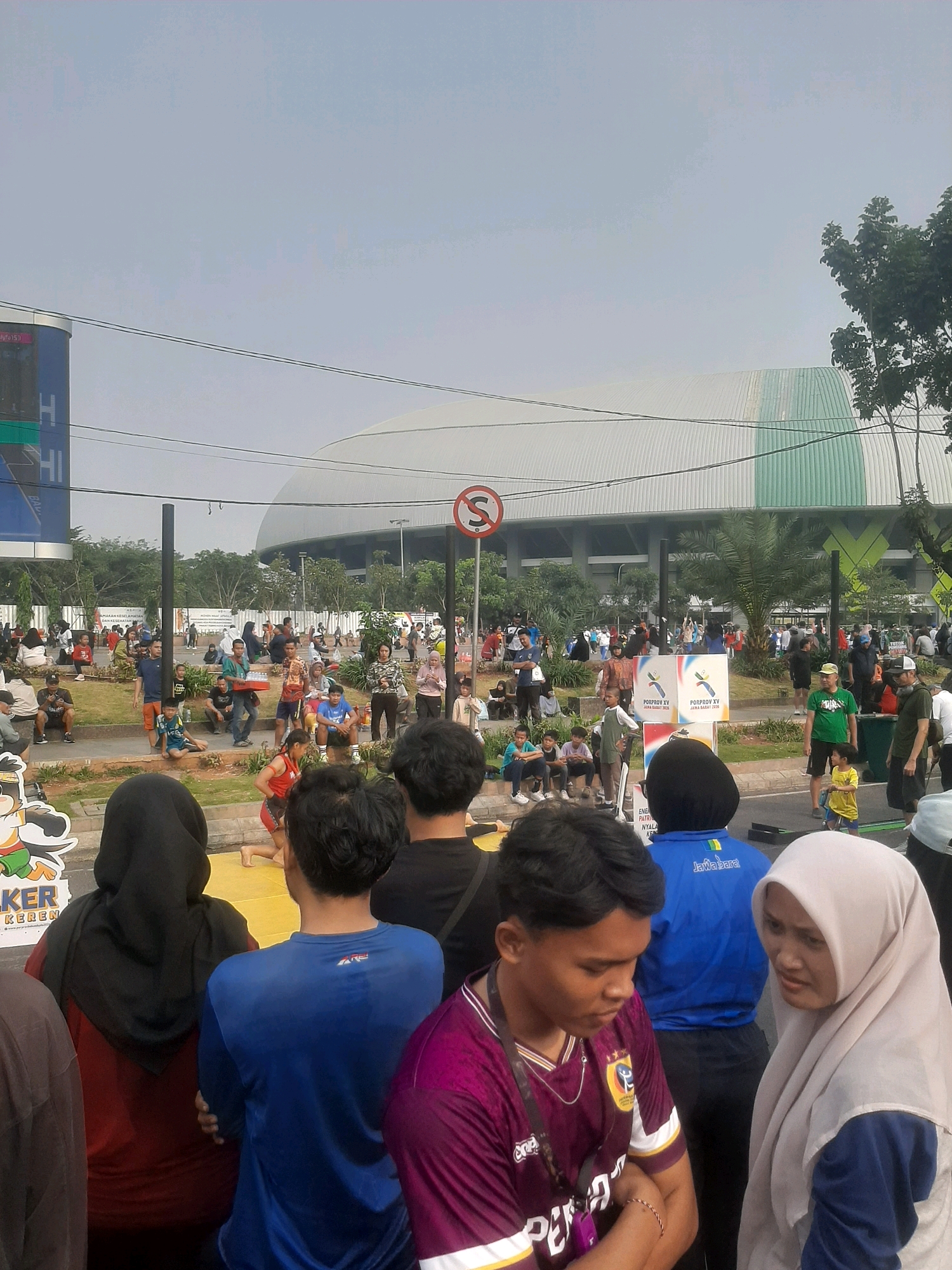 The image captures a street-level view of a gathering in front of a large, modern stadium with a distinctive white and green curved roof, likely the Patriot Candrabhaga Stadium in Bekasi, Indonesia. The sky is overcast. In the foreground, several people are visible, with a young man wearing a purple jersey with white and yellow accents in the center, looking downwards with his arms crossed. To his right, a woman in a light beige hijab is looking at him. Behind them, a person in a blue athletic shirt with "Jabar" written on the back is facing away from the camera. Further back, a diverse crowd is milling about on a grassy area and sidewalk, with some sitting and others standing. Some individuals appear to be watching an event or activity taking place on a yellow mat in the middle ground, where a person in a red outfit is visible. Several signs and banners are present, including one with a red circle and a diagonal line, indicating a prohibition, and another with logos that read "POTPROV XV JAWA BARAT 2018." The overall scene suggests an outdoor event or festival, possibly a sporting or cultural gathering.