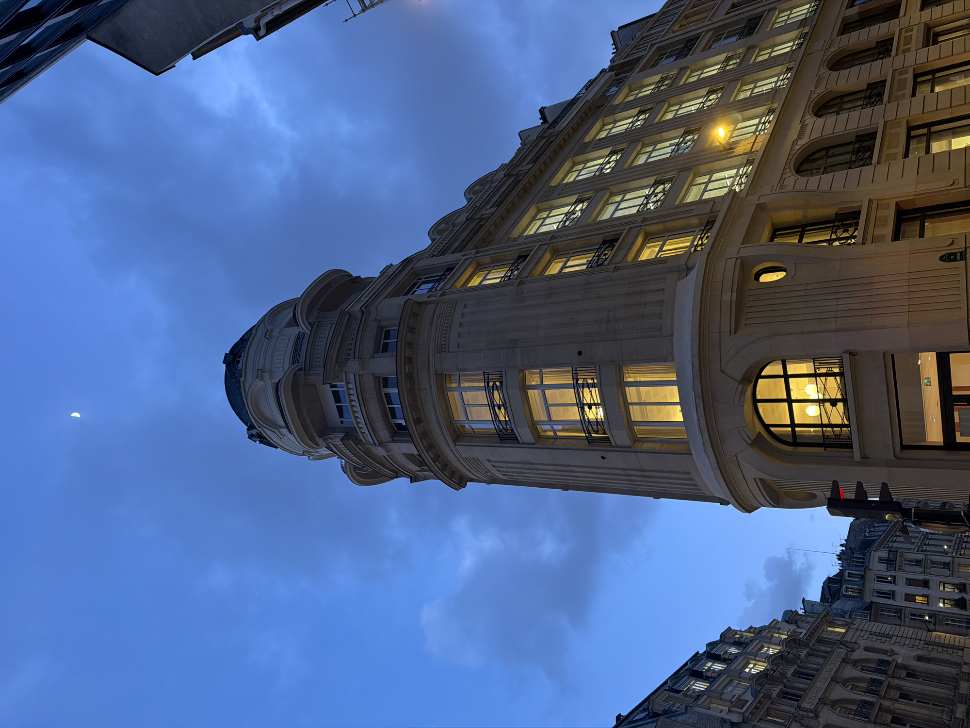 An upward-angled, wide shot captures the corner of a prominent Haussmannian building in Paris, France, at dusk or early evening. The building, identified as located at the intersection of Rue des Capucines and Place de l'Opéra (specifically 2 Place de l'Opéra / 51 Rue des Capucines), is constructed of light-colored stone with ornate classical architectural details, including pilasters, cornices, and decorative balustrades on several windows. Its most distinctive feature is a rounded corner culminating in a domed structure at the top.
Multiple windows across the building's facade are brightly illuminated from within, casting a warm yellow light that contrasts with the deep twilight blue of the sky. The sky above is partially cloudy, and a thin, white crescent moon is visible near the top center of the frame. To the immediate left of the main building, a black traffic light pole displays a red signal. Beyond it, other multi-story Haussmannian buildings line the street, also showing some illuminated windows. No people or vehicles are discernible within the visible scene. Artificial street lighting is present, with one yellow-orange streetlight illuminating the facade of a building to the right.