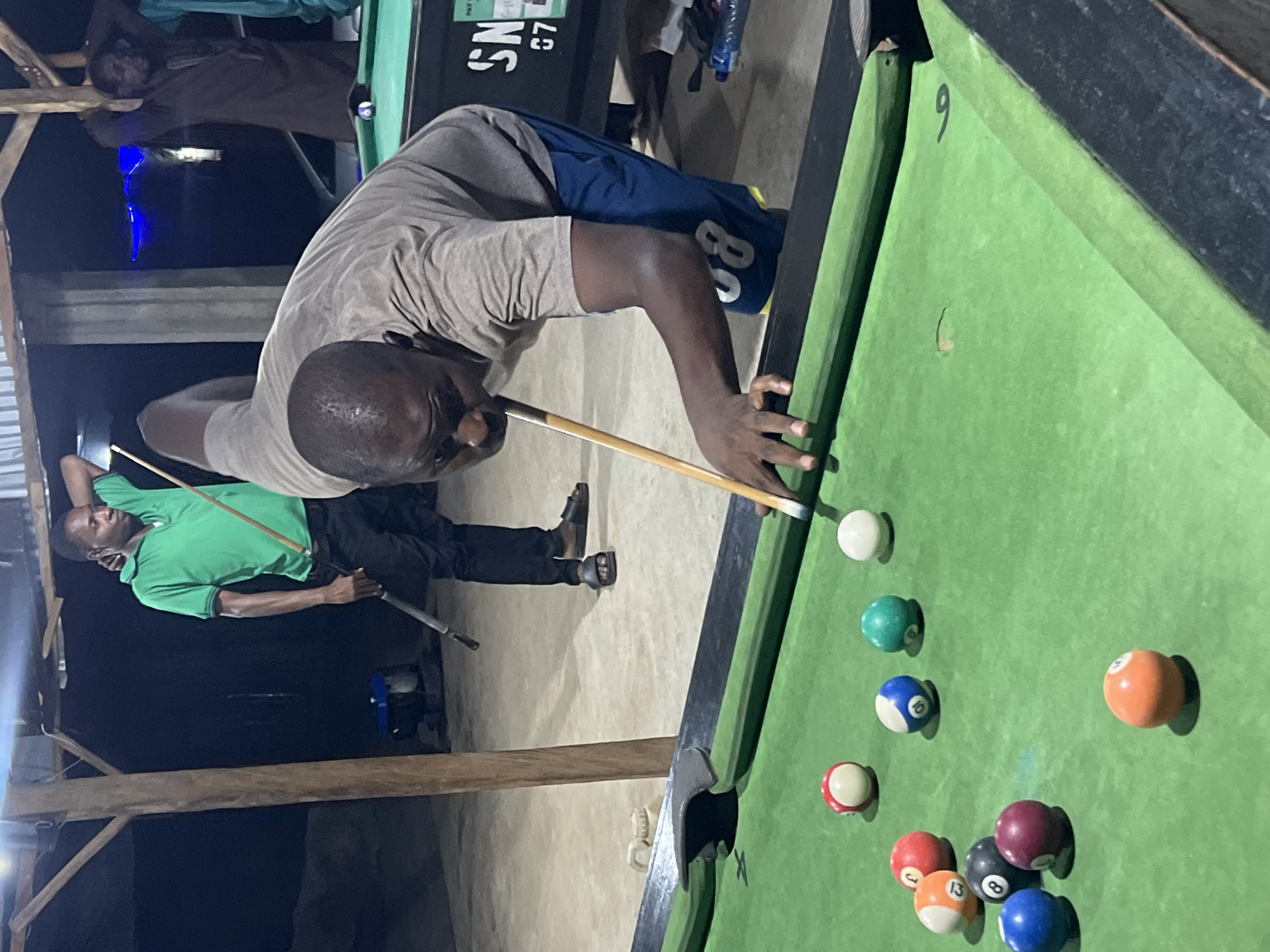 This image captures a nighttime or late evening scene in Minna, Nigeria, where men are playing pool in what appears to be a casual, semi-outdoor or indoor games area.
The central subject is a Black man, wearing a light grey t-shirt and blue shorts with a white "80" or "BO" mark, leaning intently over a green-felted pool table. He holds a wooden pool cue, his right hand forming a bridge as he aims for a shot, displaying deep concentration.
On the pool table, a white cue ball is visible, along with numerous numbered pool balls. These include solid green (6), striped blue (10), solid red (1), striped orange (13), solid black (8), striped maroon (15), solid orange (14), and solid blue (7). A number "9" is also visible on the green cushion near a pocket.
In the background, to the left, another Black man in a green collared t-shirt and dark trousers stands holding a pool cue, observing the game. A third man in a dark shirt is partially visible further back, near another pool table.
The setting features prominent wooden support beams and a dark background, suggesting artificial lighting is the primary source of illumination. In the distance, a blue-lit object, possibly a fan or light fixture, is faintly visible. On the second pool table in the background, the text "SN" and "G7" can be seen. The overall atmosphere indicates a focused yet social activity.