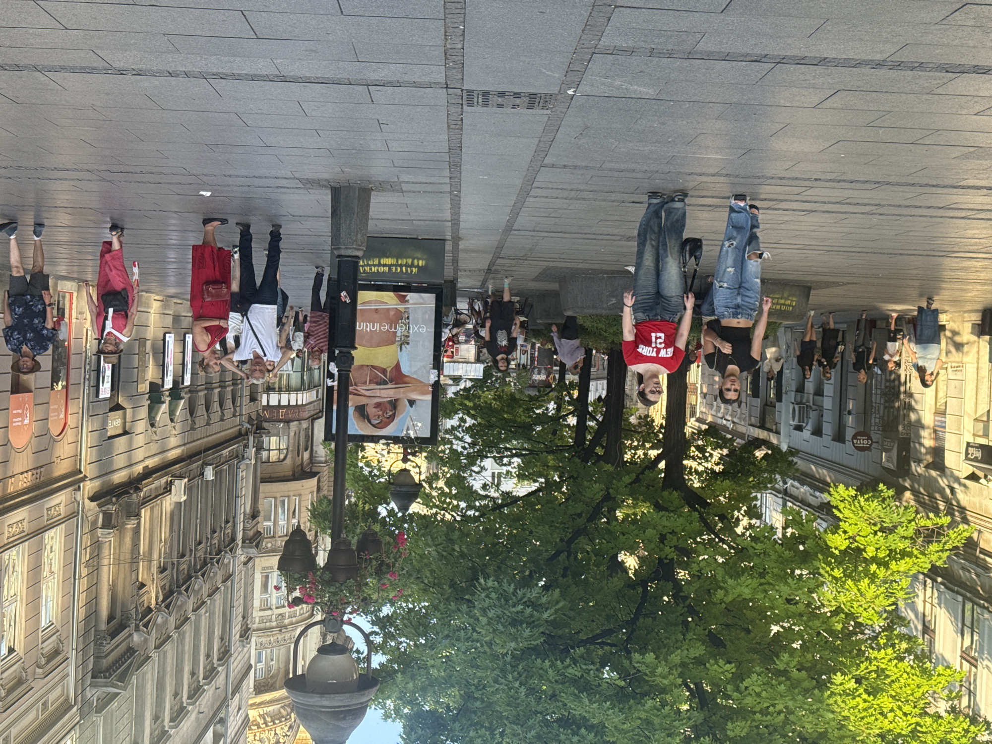 The image depicts a street scene in Belgrade, Serbia, specifically on Knez Mihailova Street. The perspective is from the street level, looking down the pedestrianized thoroughfare. Tall, ornate buildings line both sides of the street, their facades a mix of classical and art nouveau architectural styles, some with arched windows and decorative elements. Lush green trees cast dappled shadows across the wide, stone-paved pedestrian walkway. A tall lamppost with hanging flower baskets stands prominently in the mid-ground, adorned with two lamps. A large advertisement poster, featuring a woman in a hat and yellow swimwear, is visible near the lamppost.
Several individuals are walking in various directions along the street. In the foreground, two young women are walking towards the camera. The woman on the left wears a black crop top and ripped, baggy blue jeans. The woman on the right is wearing a red t-shirt with "NEW YORK 21" printed on it and similar baggy blue jeans. Further back, a diverse group of people are seen strolling, some singly, some in pairs, and some in small groups. They are dressed in casual summer attire, including shorts, t-shirts, dresses, and hats.
The lighting suggests it is late afternoon or early evening, with the sun casting long shadows and illuminating the scene with a warm glow. The environment appears to be bustling with activity, indicative of a popular urban public space. The overall impression is one of a vibrant European city street on a pleasant day.