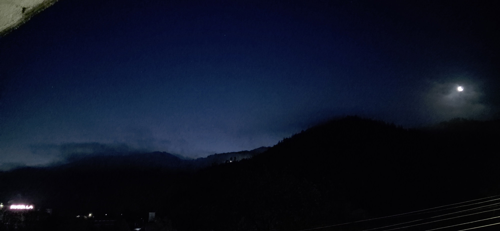 The image depicts a nighttime view in Rudozem, Bulgaria, showing silhouetted mountains under a dark blue sky. The moon is visible through scattered clouds in the upper right. In the foreground and to the left, faint lights of the city are discernible. Power lines are visible in the bottom right corner. The scene is dark and indistinct.