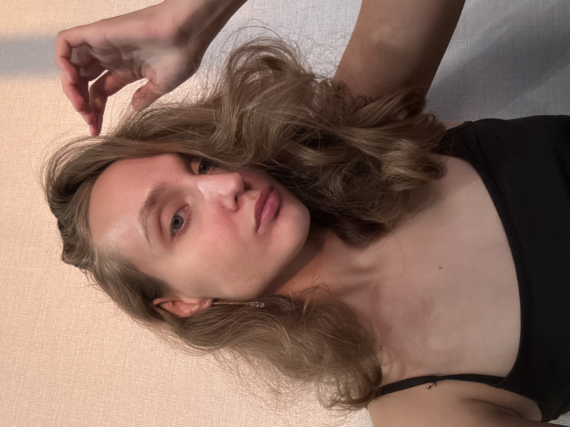 The image is a close-up, waist-up selfie of a young woman with long, wavy, light brown hair. She is wearing a black tank top and a delicate gold earring with a small charm. Her right hand is raised, touching her hair near her temple. She has light blue eyes, fair skin with some redness on her cheeks, and pale pink lips. The background appears to be a textured, light-colored wall. There is no discernible information within the image that suggests a specific city or location, including Подшивалово, Russia.