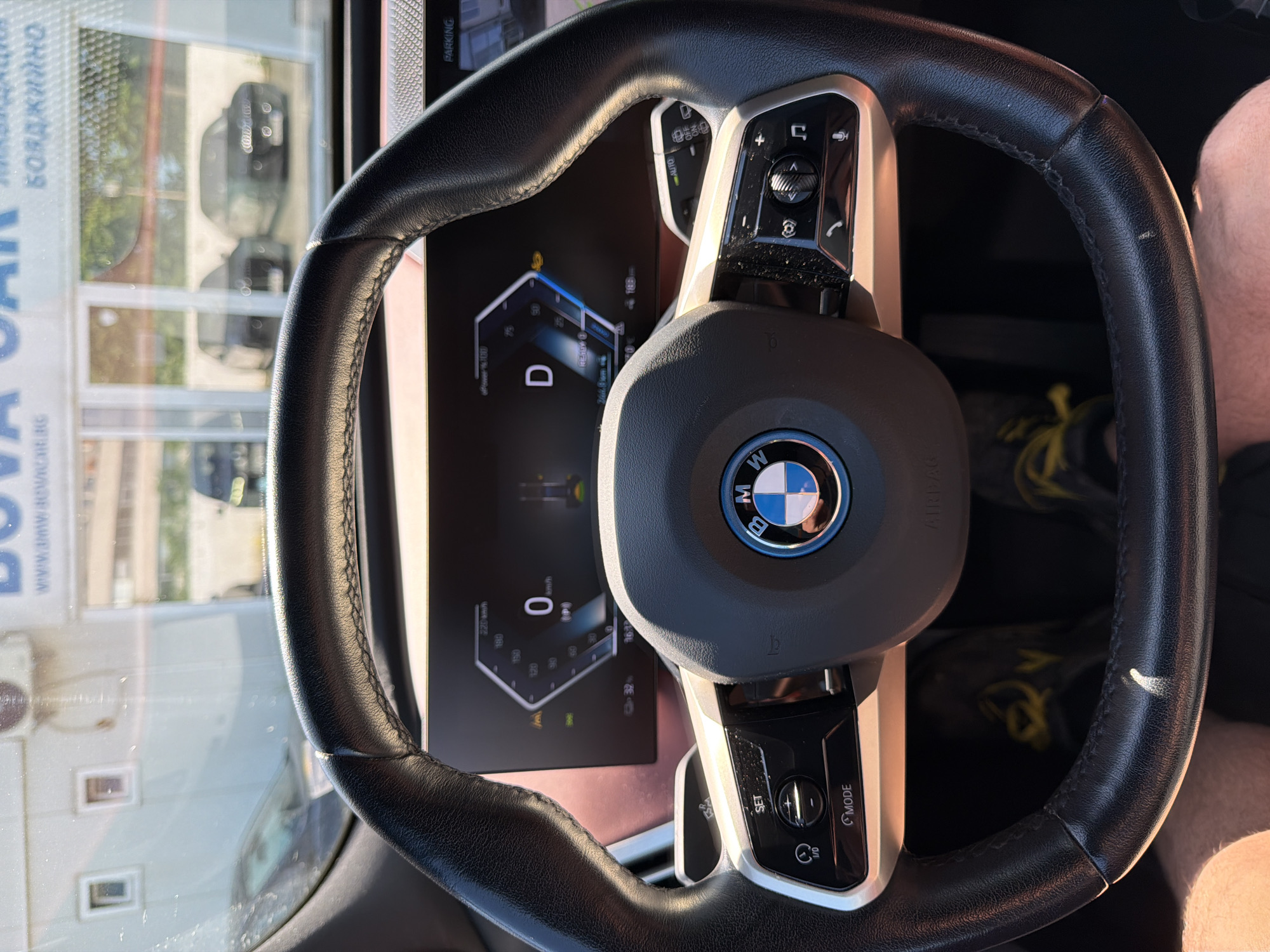 The image shows a close-up view of the interior of a BMW car, likely a modern model given the digital dashboard. The perspective is from the driver's seat, looking at the steering wheel and the dashboard display. The steering wheel is black leather with silver accents and features the BMW logo in the center. Various buttons are visible on the steering wheel, including controls for cruise control ("SET", "MODE") and media. The digital dashboard displays a speed of "0 km/h" and "D" for drive. The time "16:32" and a distance of "244.8 km" are also visible on the screen. The background shows a reflection in the car's windshield, revealing an outdoor environment with buildings and other vehicles. Through the windshield, two dark-colored cars are visible parked outside. Above the steering wheel, a sign with text that appears to be "BOVA CAR" and partially visible Russian text "БОЯДЖИИНО" is reflected. This signage, combined with the use of the Cyrillic alphabet and the specific make of car, suggests the location is likely a car dealership or service center in Burgas, Bulgaria, which uses both Latin and Cyrillic scripts in its signage. The overall lighting suggests it is daytime. A person's legs wearing black shoes with yellow accents are visible in the lower part of the frame, indicating the presence of the driver.