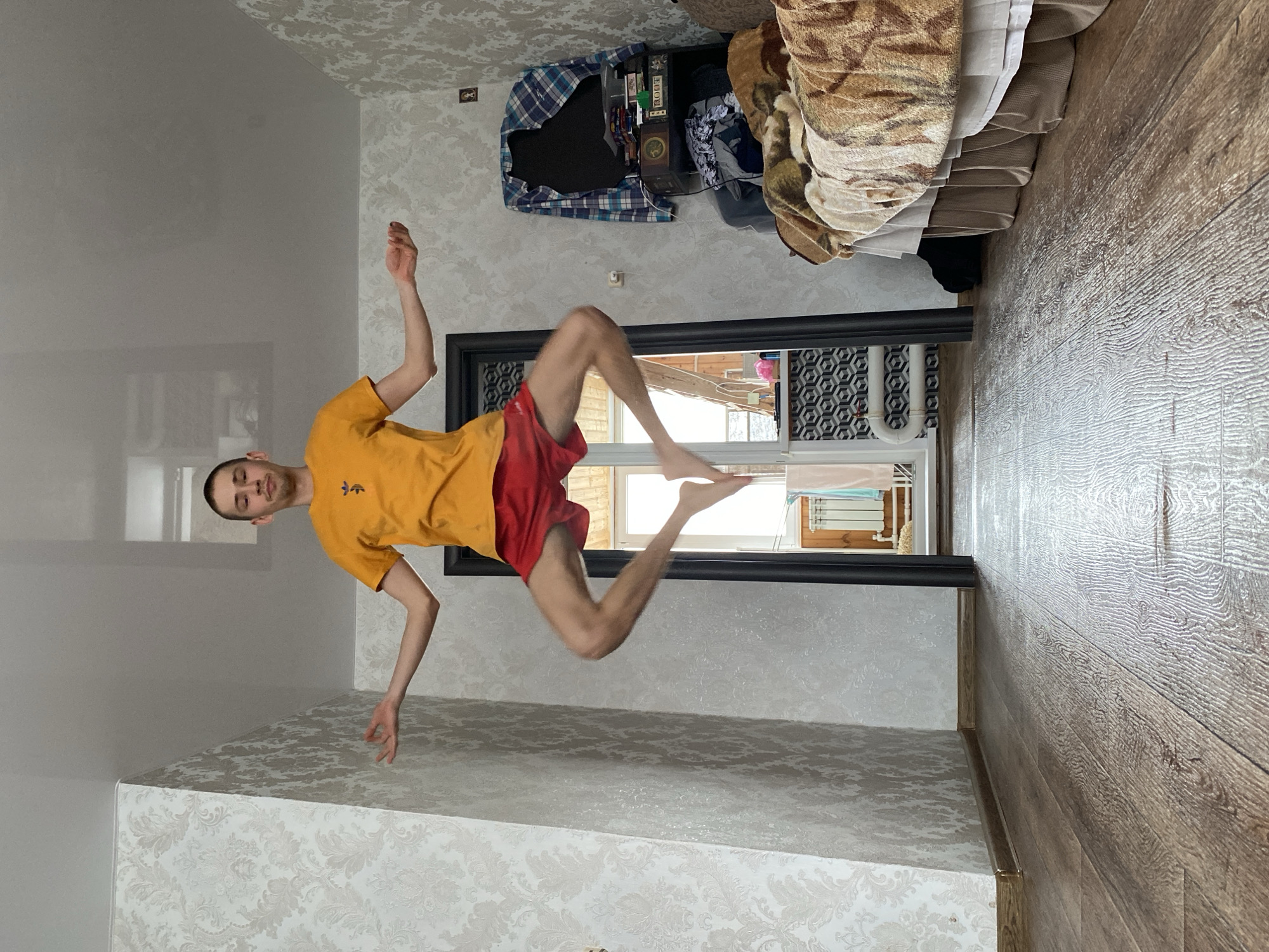 An indoor scene in what appears to be a room or apartment in Ulyanovsk, Russia, features a young man in mid-air. He is light-skinned, with short dark hair, wearing a bright yellow t-shirt with the "adidas" trefoil logo and red shorts. His arms are bent and extended outwards, and his legs are drawn up, suggesting a jump or levitation. He looks towards the viewer with a calm expression.
The room has light-colored patterned wallpaper on two visible walls, and a plain light gray wall towards the back. The floor is covered with wooden laminate. A large window with a black frame is visible on the plain wall, letting in natural daylight. Underneath the window, a white radiator is partially visible.
To the right side of the image, a bed or sofa is partially visible, covered with textured blankets in brown and beige tones, possibly with an animal print. Above the bed/sofa, a dark shelf or dresser holds various items, including a plaid shirt, several books (one clearly labeled "DOOM"), and other personal belongings. The overall atmosphere is casual and domestic, with the man's dynamic pose providing a focal point. The time of day appears to be daytime due to the light from the window.