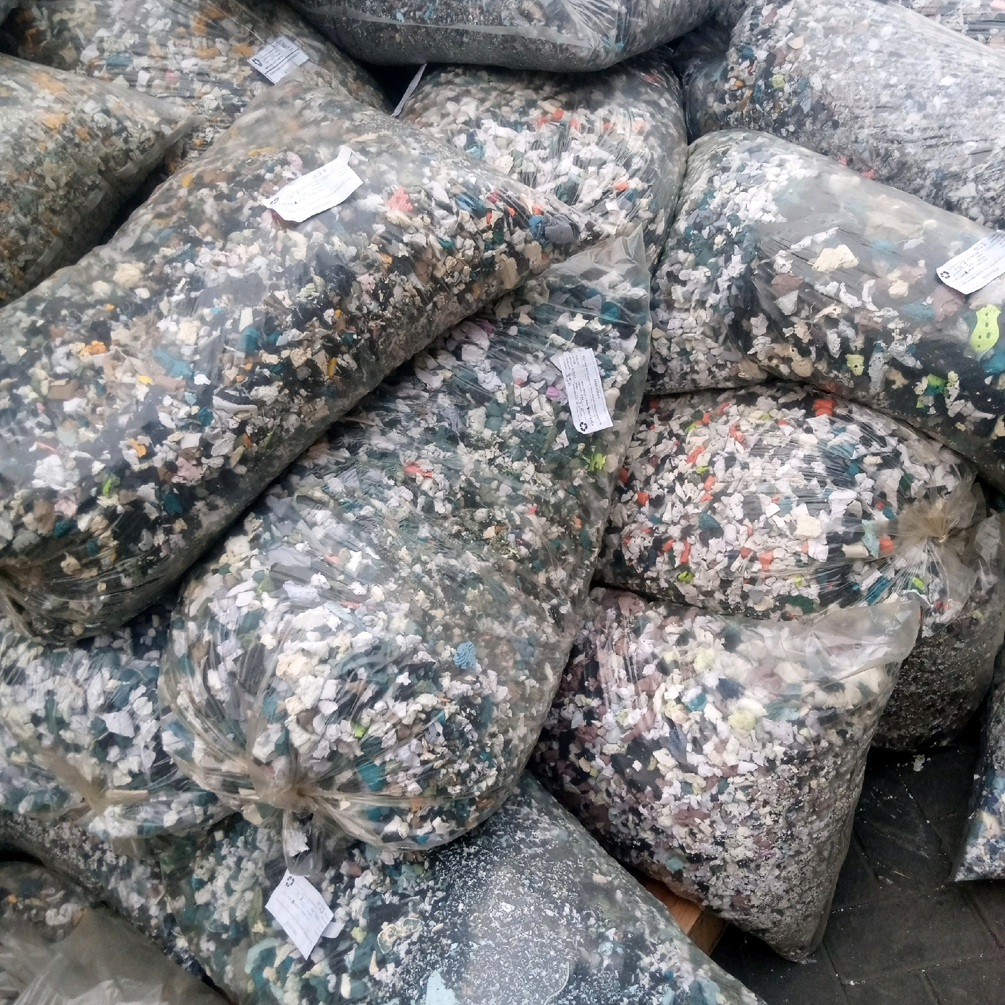 The image shows a close-up view of several large, clear plastic bags filled with shredded foam or similar material. The material inside the bags is multi-colored, with visible pieces of white, grey, blue, green, and pink. Some of the bags have white labels attached to them, which appear to be product information or recycling symbols. The bags are piled together, some on top of others, and they are resting on a dark, possibly concrete surface. The lighting suggests an outdoor or industrial setting. There are no people visible in the image. The image appears to have been taken in Cijeruk, Indonesia.