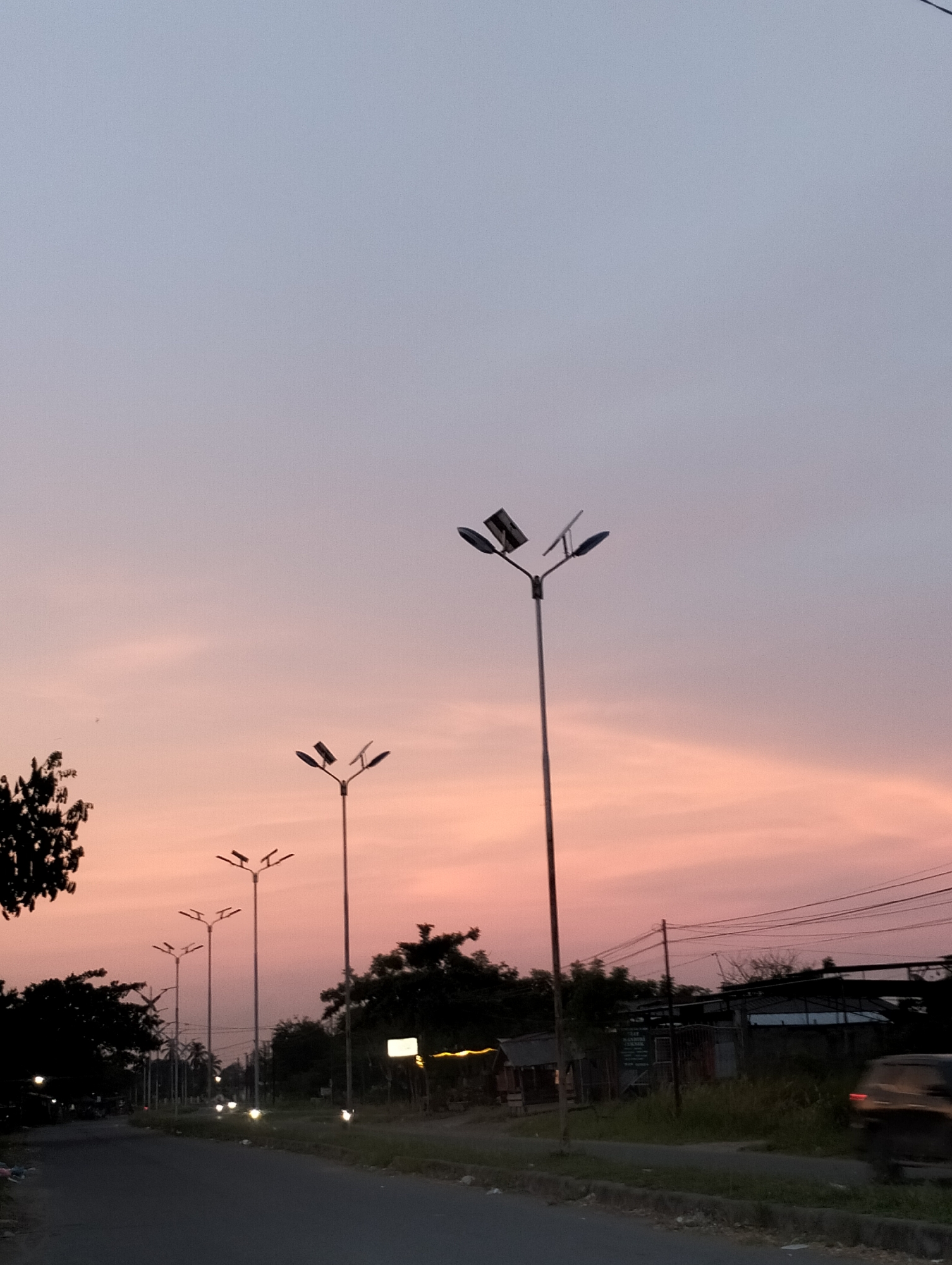 This vertical image captures a serene road scene during sunset or dusk in Lhokseumawe, Indonesia. The sky occupies the majority of the frame, displaying a striking color gradient from a soft lavender-blue at the top, fading into warm pinks and oranges closer to the horizon, with subtle, wispy cloud patterns.
A line of tall streetlights with multiple fixtures extends along the right side of the road, receding into the distance. The closest prominent streetlight, slightly right of center, features a main pole with several arms, each equipped with what appears to be a flat rectangular panel, possibly a solar panel or reflector, along with a light fixture. The ground and the road are significantly darker, enveloped in shadow, which creates a strong contrast with the vibrant sky.
On both sides of the road, dark silhouettes of trees and dense foliage are visible, with a large tree partially seen on the far left. On the right, behind the streetlights and trees, the dark outlines of small structures, possibly sheds or simple buildings with corrugated roofs, can be vaguely discerned. Overhead, a network of electrical wires crisscrosses against the twilight sky. A blurred, dark-colored vehicle is visible on the far right, suggesting movement on the road. The overall mood is peaceful and tranquil, characteristic of the transition from day to night. No discernible text is visible on any objects or signs within the image.