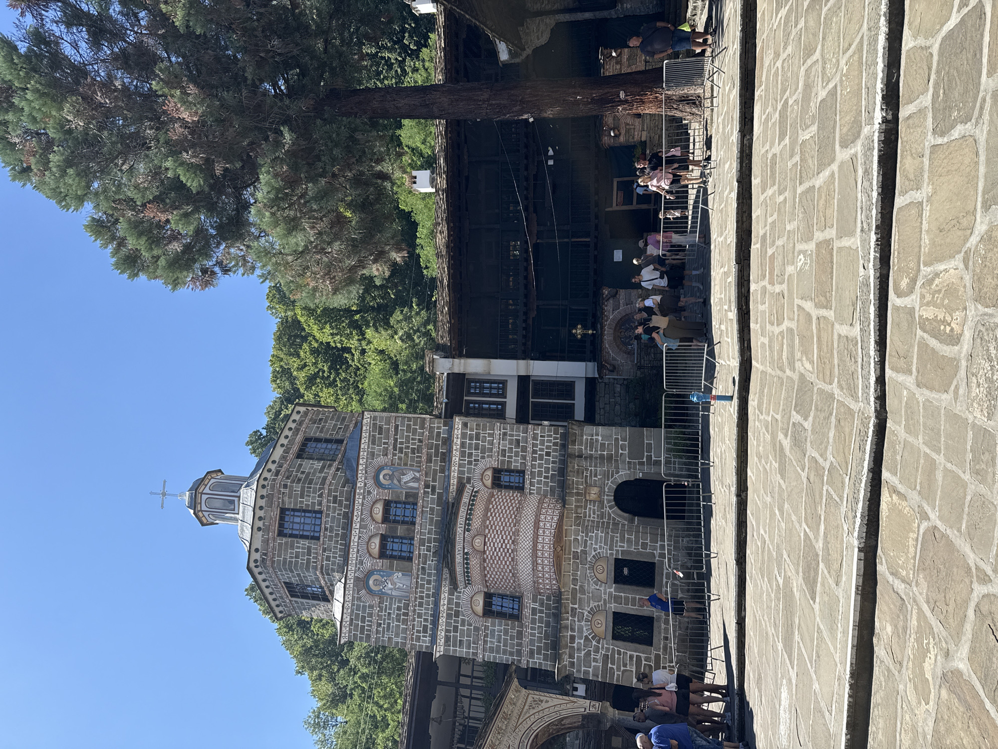 This image shows the exterior of the Rila Monastery in Oreshak, Bulgaria, on a sunny day. The monastery is a large, historic complex with stone architecture. In the foreground, a courtyard paved with large flagstones is visible, with a raised section in the center. A metal fence separates the courtyard from the buildings and a group of tourists.
The main structure in the image is a multi-story stone building with a distinctive octagonal tower topped by a dome and a cross. The tower features arched windows, decorative brickwork, and religious frescoes. Adjacent to this building is a long, dark wooden structure with balconies and a sloped, tiled roof. A large, mature tree with green and brown foliage stands to the right of the main building, partially obscuring the view of the wooden structure and the surrounding trees.
Several people are standing in the courtyard, some looking towards the monastery, others gathered in groups. They are dressed in casual summer clothing. A blue fire hydrant is visible near the fence. The background is filled with lush green trees, indicating a mountainous or forested environment surrounding the monastery. The sky is clear and blue.