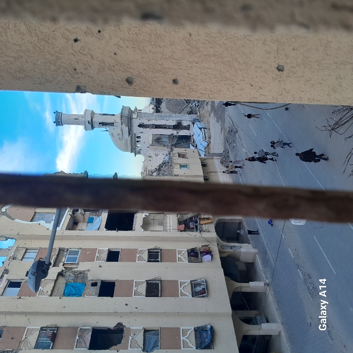 A damaged building with missing windows faces a partially destroyed mosque. People walk along the adjacent street. The buildings show significant damage, possibly from conflict. A metal bar partially obscures the view. A blue sky is visible above the mosque. The overall atmosphere is one of devastation and resilience.