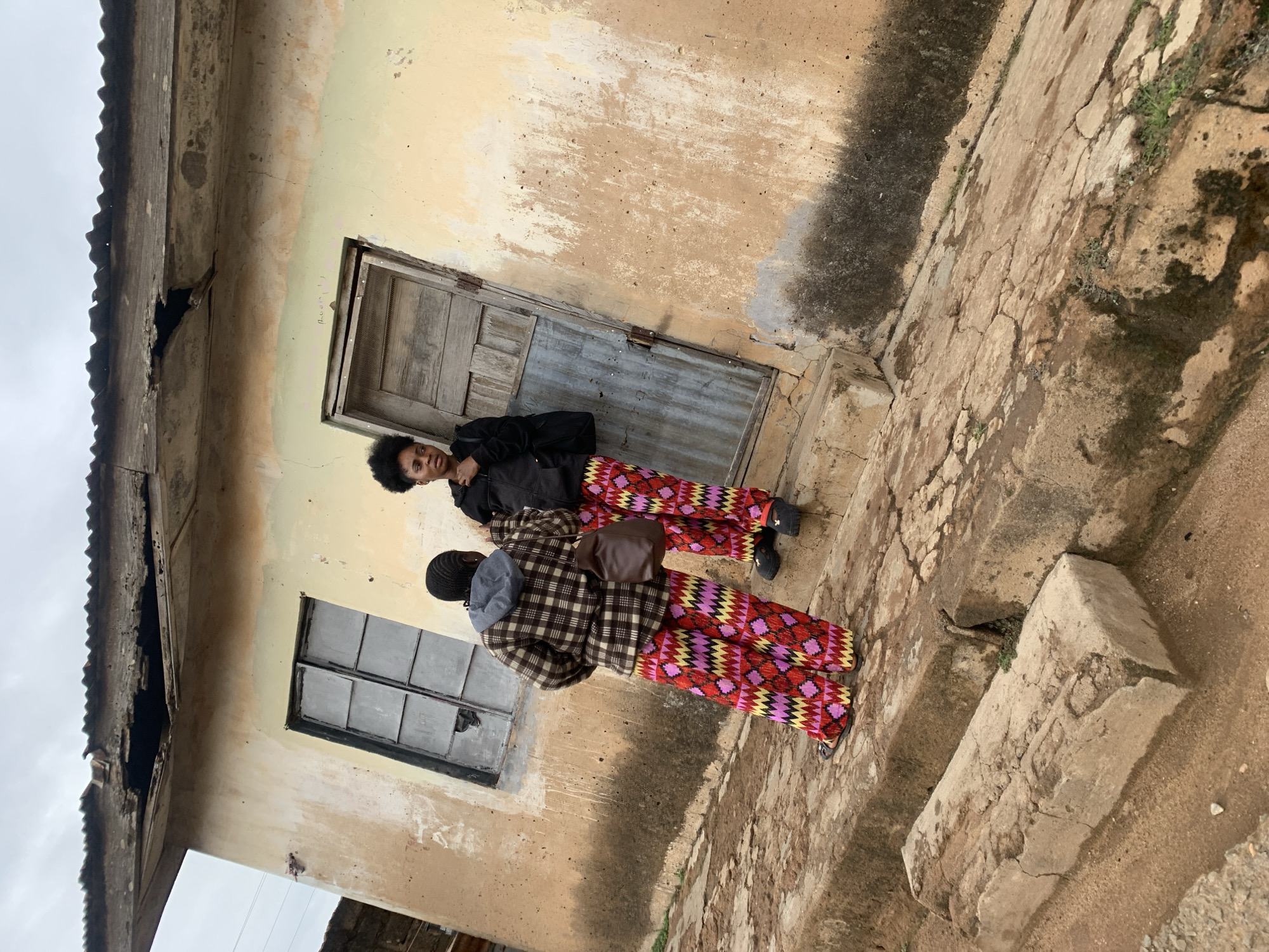The scene depicts two individuals standing next to a deteriorated building in Jos, Nigeria. One person is wearing a black jacket and patterned trousers, while the other is wearing a plaid jacket and patterned trousers. The building has a damaged roof and weathered walls with barred windows and a wooden door. The ground is paved with cracked stone slabs, some overgrown with vegetation.
