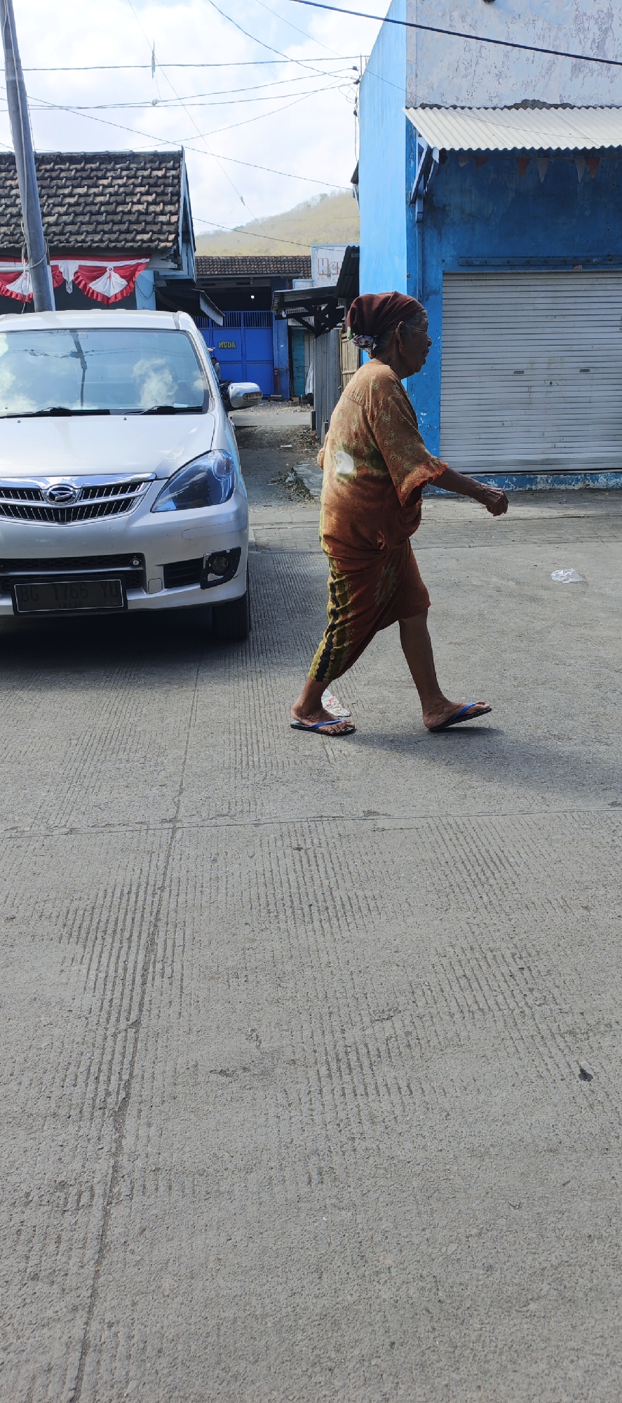 An elderly woman with a brown headscarf and an orange and brown patterned sarong is walking on a paved street in Puger, Indonesia. She is wearing blue flip-flops. To her left is a silver minivan. Behind her, on the right, is a blue building with a corrugated metal door and a white awning. On the left, there is a building with a red and white striped banner. The sky is partly cloudy.