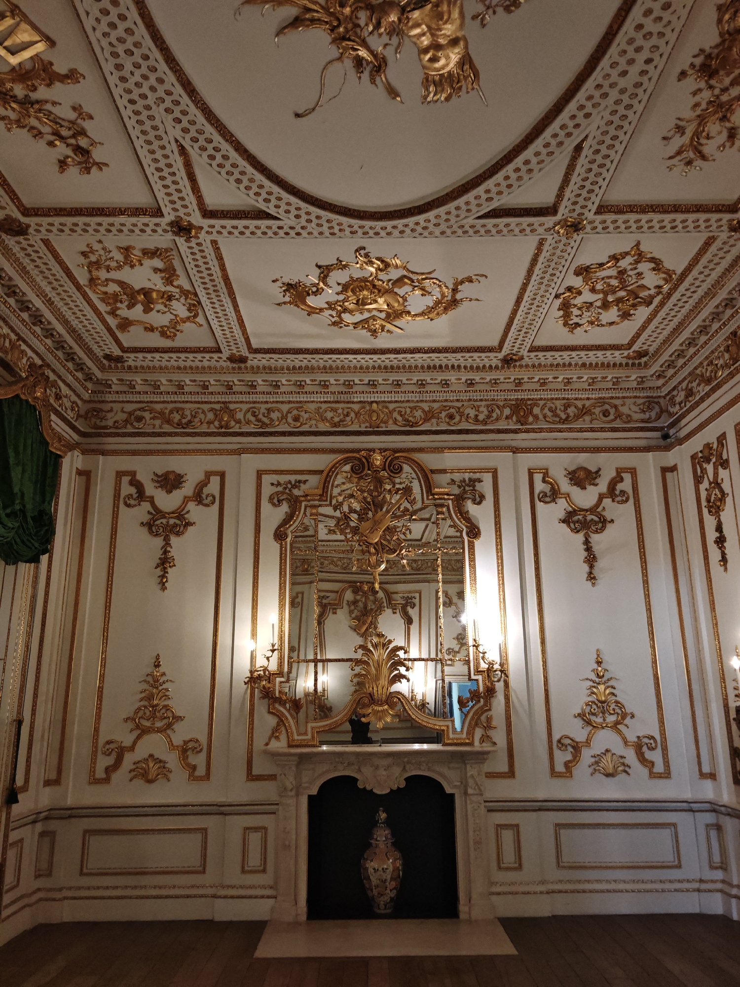 The image depicts an ornate interior room with a high ceiling featuring intricate gold detailing and a large, decorative central fixture. The walls are panelled and adorned with gold-trimmed mouldings and decorative motifs. A marble fireplace with a dark hearth is flanked by candelabras and topped by an elaborate gold embellishment that extends upwards, incorporating a mirror. A large vase sits within the fireplace. There is a green curtain visible on the left. The room appears to be within a historical building. Although the location in the city of London, United Kingdom is mentioned, there is no information given in this image that supports that assertion.