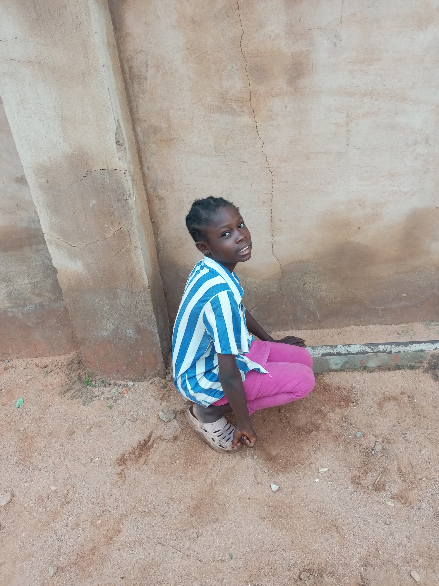 A young girl with dark skin and braided hair is crouching down in front of a beige, cracked, concrete wall. She is wearing a blue and white striped t-shirt and bright pink pants. Her feet are covered in beige sandals. The ground is sandy with scattered pebbles and a few small green plants. The wall appears weathered, with visible cracks and discoloration. The light suggests it is daytime, possibly late morning or early afternoon. The girl is looking towards the camera with a neutral expression. The scene appears to be outdoors, in a dusty environment, consistent with a location in Tempure, Nigeria.