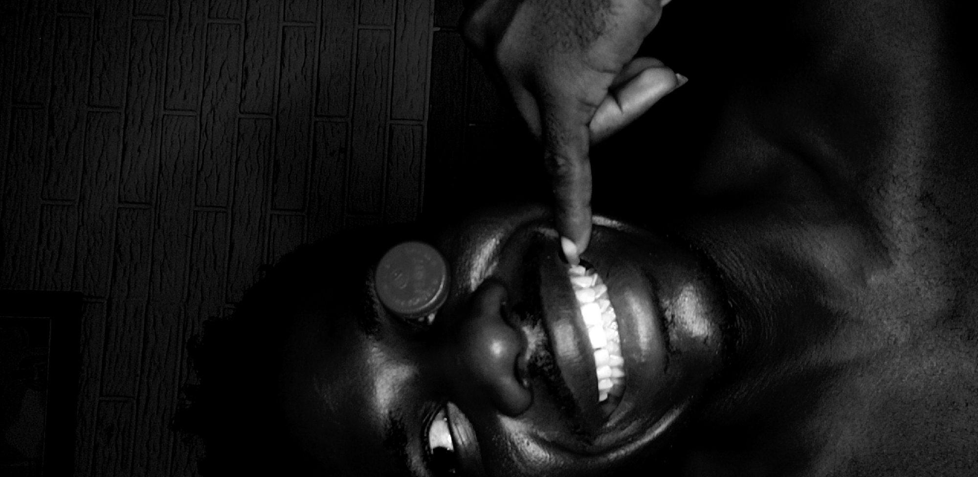 This black and white, high-contrast close-up image features a person's face and a hand against a dark, textured background. The subject's face, with dark skin, is viewed from a slightly overhead perspective, tilted such that the right side is more prominent, with eyes looking upwards. Their mouth is open, revealing a row of distinctly white teeth.
A coin, resembling a Nigerian 10 Naira coin, is balanced on the person's forehead or temple area, with faint circular text and numbers visible on its surface. Another person's hand, also with dark skin, extends into the frame from the top. The index finger of this hand is positioned inside the mouth, gently touching the upper front teeth. The fingernail on the index finger is visible.
The background is a dark, vertically aligned surface with a discernible pattern of rectangular blocks or planks, suggesting a wall texture. The lighting is dramatic, casting deep shadows and highlighting the brighter elements like the teeth and fingernail. The scene appears staged, capturing an intimate and focused interaction, possibly playful or symbolic. No explicit emotions are clearly depicted, but a sense of concentration or calm prevails. The faint text on the coin is not clearly legible.