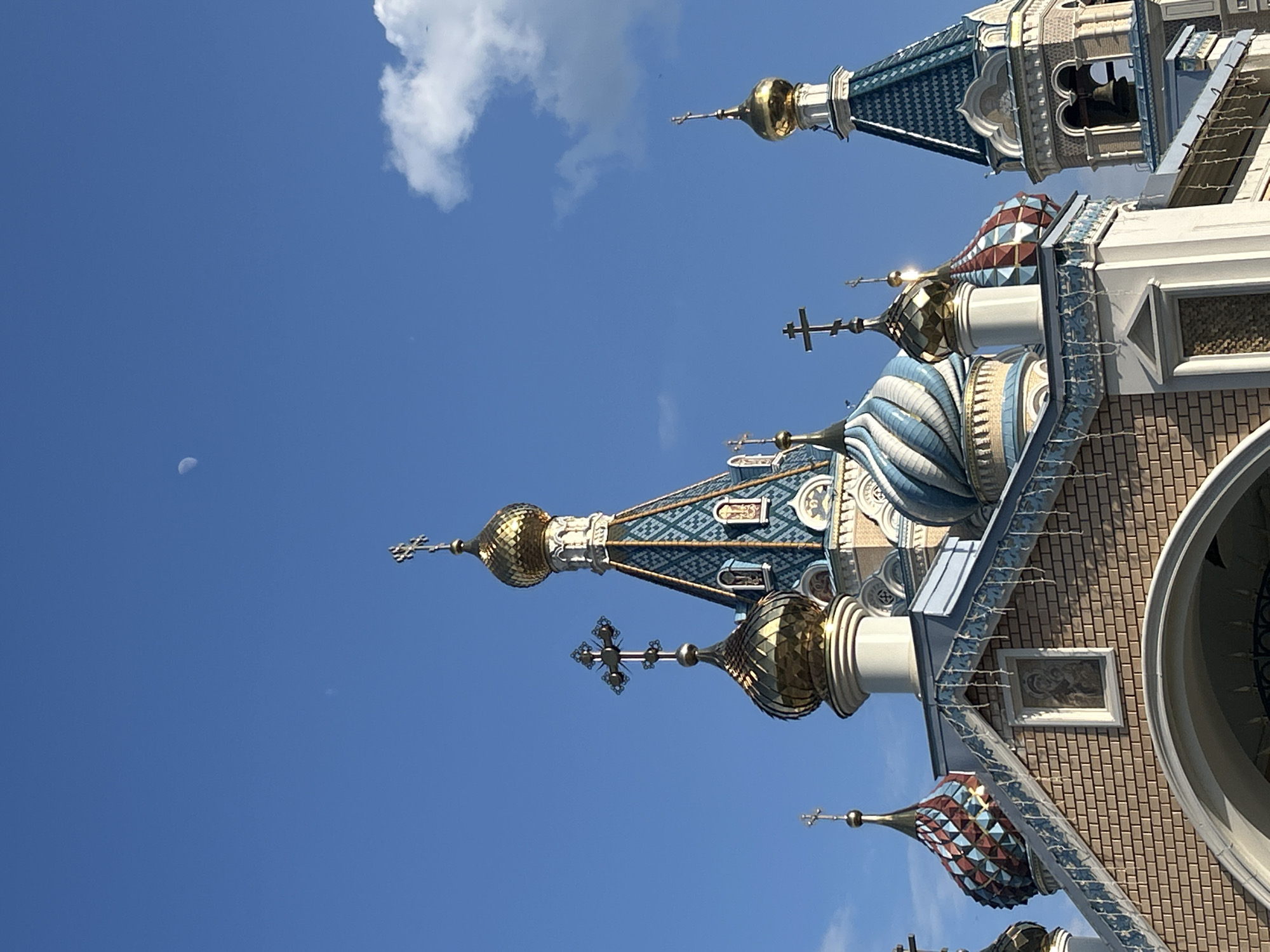 The image shows the exterior of St. Michael's Cathedral in Izhevsk, Russia, with a blue sky as a background. The view is from below, capturing the intricate details of the cathedral's domes and architectural elements. The domes are adorned with colorful geometric patterns and topped with golden crosses. There are a few clouds in the sky, and a sliver of the moon is visible on the left side of the image. The cathedral's brick facade is partially visible on the right.
