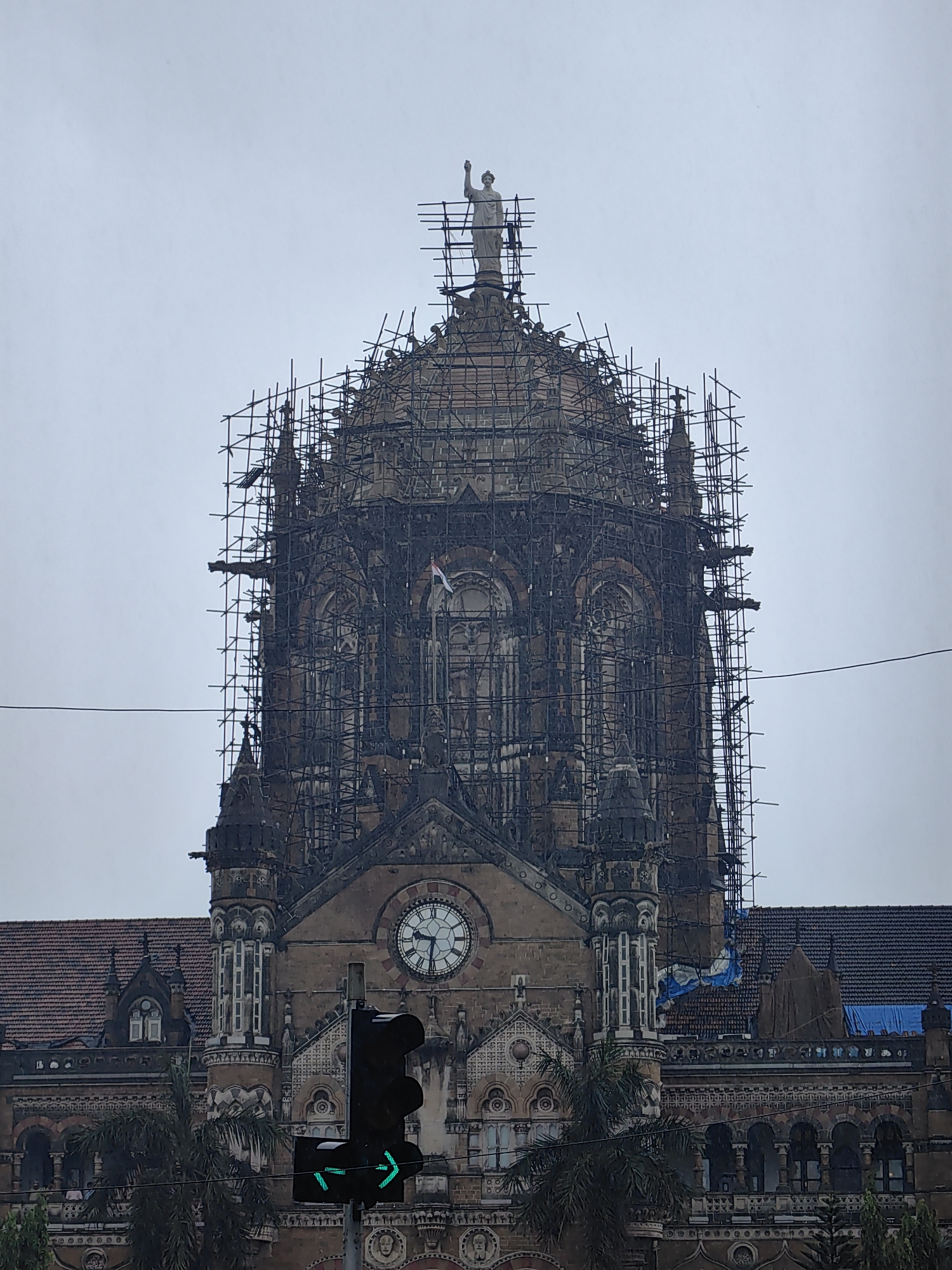 The image shows a close-up, eye-level, exterior shot of the upper portion of the Chhatrapati Shivaji Maharaj Terminus (CSMT) in Mumbai, India, on an overcast day. The iconic building's central dome and facade are covered in extensive scaffolding, suggesting ongoing renovation or maintenance work. A statue of a winged angel stands atop the dome. The detailed architectural features of the building, including arches, windows, and decorative elements, are visible beneath the scaffolding. A large clock is prominently displayed on the facade. In the foreground, a traffic light shows a green arrow pointing to the right, and a green light is illuminated. Palm trees are partially visible in the lower part of the frame, indicating a tropical or subtropical environment. The overall impression is of a historic landmark undergoing preservation.