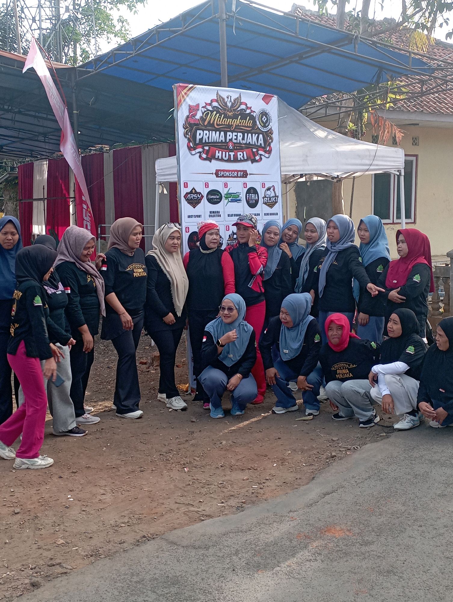 The image depicts a group of women, likely participants in an event, gathered outdoors. A banner in the background features text in Indonesian, including "Milangkala PRIMA PERJAK & HUT RI," which translates to "Anniversary PRIMA PERJAK & Republic of Indonesia's Independence Day." This suggests the event is a celebration of an anniversary and Indonesia's Independence Day. Several women are wearing matching black tops with the same text printed on them, indicating they belong to a group or organization. Some are standing, while others are crouching or kneeling. They are all wearing headscarves, common attire in Indonesia. The environment appears to be a somewhat informal outdoor setting, possibly a street or a communal area, with a tent structure and a building visible in the background. The presence of banners and a gathering of people suggests a local community event. The women are diverse in age and expression. The ground is a mix of dirt and gravel, with some paved road visible. The overall scene is one of communal gathering and celebration, likely in a public space within Tasikmalaya, Indonesia.