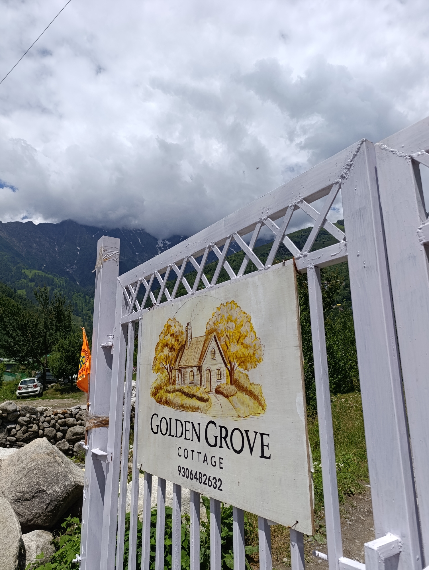 The image captures the entrance to "Golden Grove Cottage" in Barheta, India, on a cloudy day. A white wooden gate, adorned with a patterned lattice, stands open, revealing a sign with a painted cottage and trees. The sign reads "GOLDEN GROVE COTTAGE" in black text, followed by the phone number "9306482632". In the background, a white car is parked near a stone wall, and beyond that, a verdant hillside slopes up to majestic, cloud-shrouded mountains with snow-capped peaks. An orange flag with a yellow symbol flutters on a pole near the entrance. The foreground shows large rocks and green foliage.