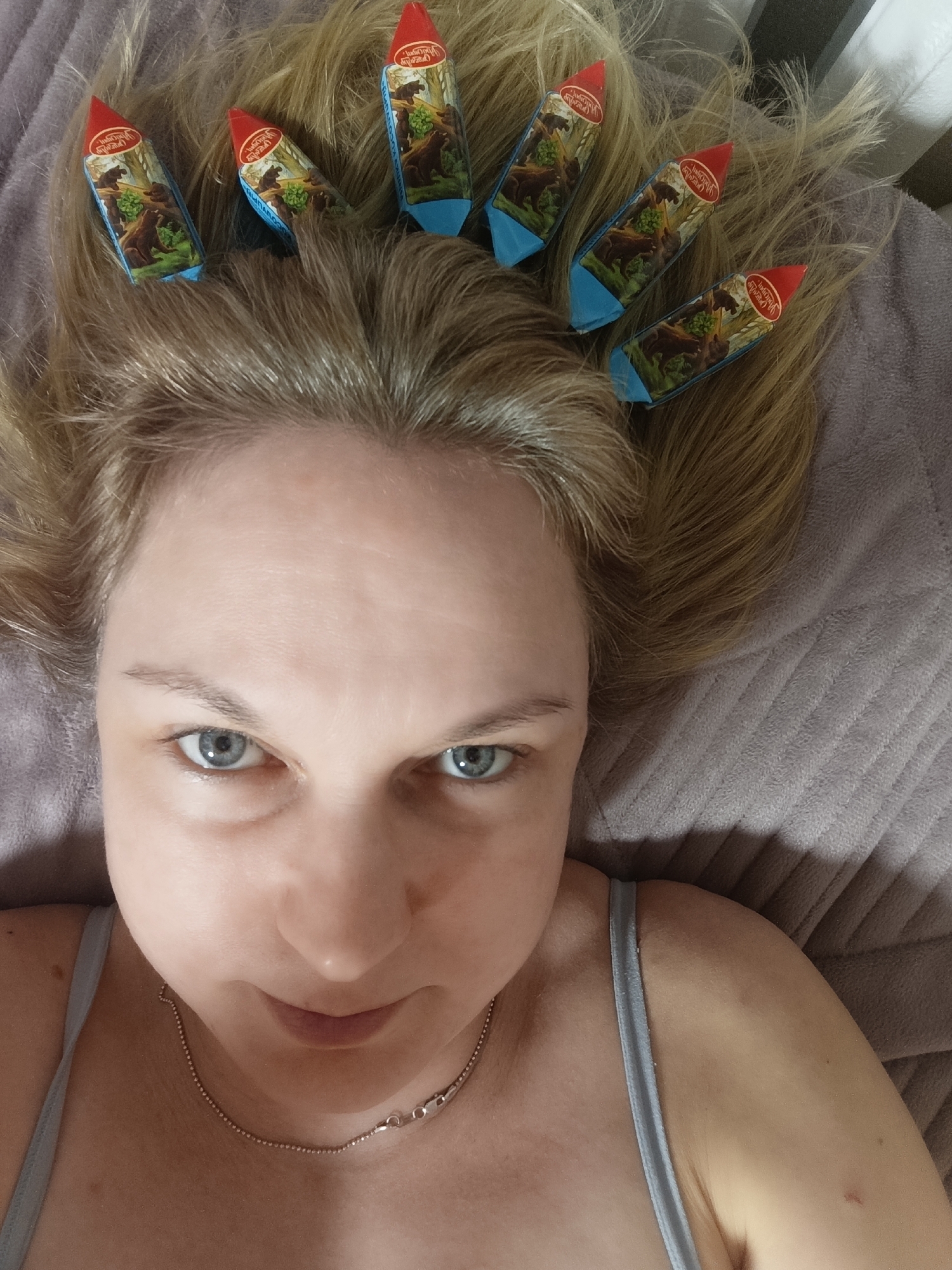 A woman with fair skin, blue eyes, and light brown hair is lying on a light purple, textured surface, possibly a bed or couch. She is wearing a light-colored tank top and a thin silver chain necklace. Six small, wrapped cheese wedges with a red top and blue and brown packaging are placed in her hair across her forehead. The packaging features an image of bears in a forest. The photograph is taken from a slightly overhead perspective, focusing on the woman's face and the cheese wedges in her hair. The location is Марковский, Russia.