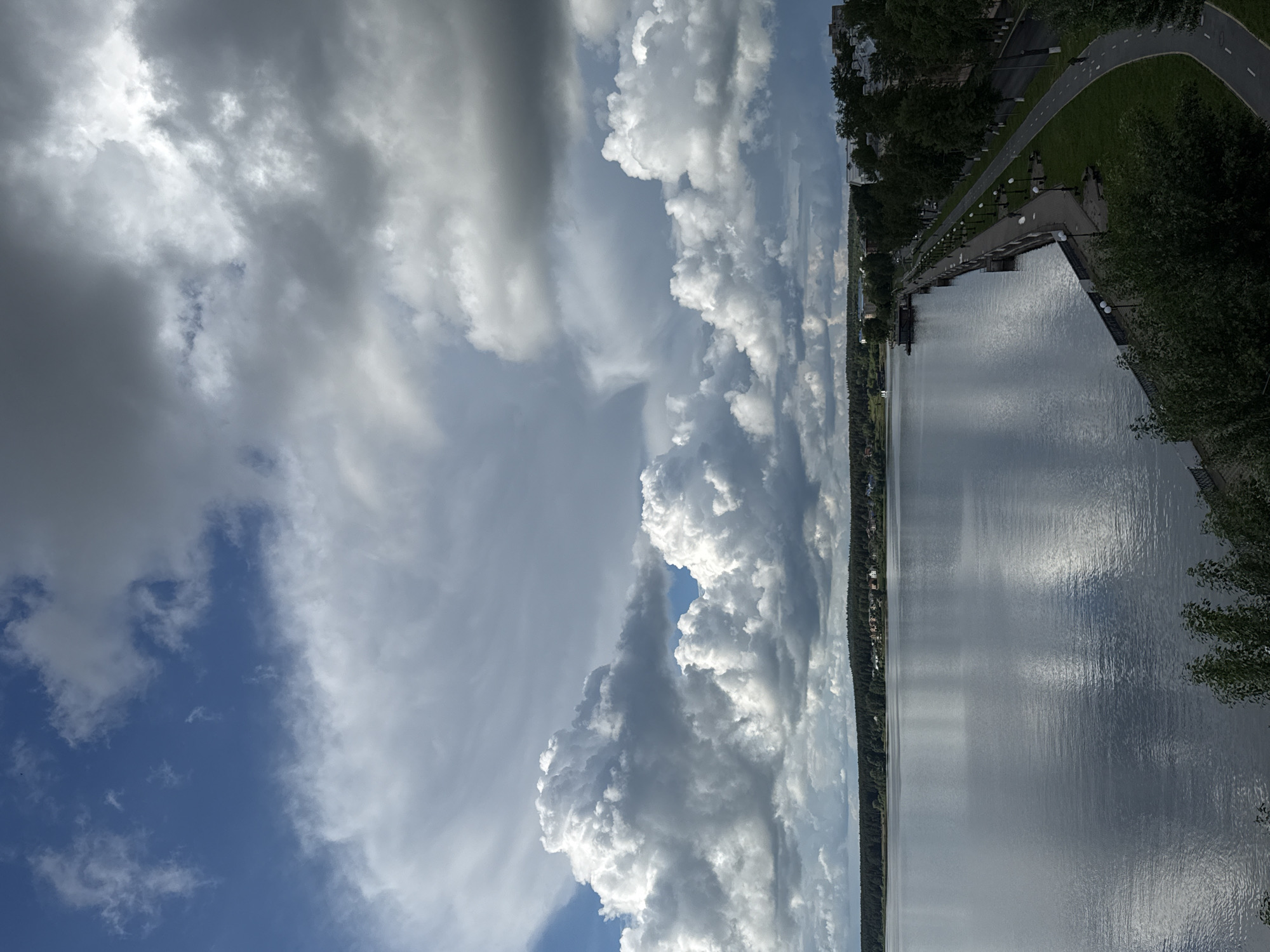 The image displays a panoramic landscape featuring a large body of water, an expansive sky, and a developed shoreline. The perspective is from an elevated vantage point, looking across the water.
The sky occupies a significant portion of the scene, filled with a diverse array of clouds. Bright, towering cumulus clouds are prominent, especially on the right and in the lower center, illuminated by sunlight. These contrast with large, darker grey cloud masses and patches of lighter, diffuse clouds, while areas of clear blue sky are visible, particularly towards the left.
The calm surface of the water, which appears to be a lake or wide river, stretches horizontally across the middle of the frame. It beautifully reflects the varying cloud patterns and the bright sunlight, creating shimmering streaks and broad reflections.
Along the right side of the image, an urban embankment runs parallel to the water. It features a paved pathway, likely a promenade or bike lane, marked with light lines. This path is bordered by well-maintained green grass and numerous trees, suggesting a park-like environment. A series of regularly spaced lampposts or streetlights are visible along the water's edge of the path. Further back from the shore, partially obscured by trees, some buildings can be seen, indicating an urban or suburban setting.
The distant left bank of the water body is characterized by a dense line of dark green trees, forming a forested horizon, with faint hints of structures or a settlement in the distance.
No people or specific activities are depicted, conveying a sense of tranquility and vastness. The scene captures daytime conditions with dynamic cloud cover, suggesting either changeable weather or the progression of a sunny day with developing clouds. There is no visible text in the image. The location is contextualized as being in the vicinity of Votkinsk, Russia.
