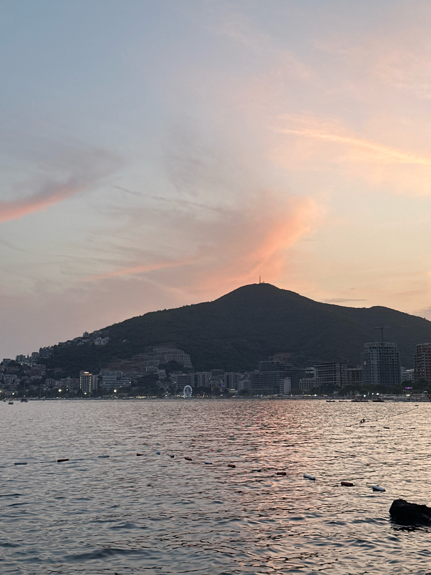 The image depicts a coastal city scene at sunset. A dark, tree-covered mountain dominates the background, with a radio tower visible at its summit. The slopes of the mountain are dotted with buildings, suggesting a densely populated hillside. In the foreground, a body of water, likely the Adriatic Sea, stretches across the frame. The water is choppy and reflects the soft, pastel colors of the sunset sky, which is adorned with streaks of pink and orange clouds.
Along the coastline, a collection of modern buildings, including hotels and apartments, form a prominent silhouette against the darkening mountain. Some buildings are illuminated with lights, indicating it is twilight. A Ferris wheel, lit up with white lights, is visible in the distance on the right side of the cityscape.
In the water, a series of buoys or markers are visible, forming a line that separates an area of the sea. The water surface is textured by ripples and waves. A dark rock is visible in the lower right corner of the image, partially submerged in the water. No people are clearly discernible in the image, but the presence of buildings and lights suggests human activity. The overall impression is one of a tranquil evening scene in a coastal resort town. The specific location is Budva, Montenegro.