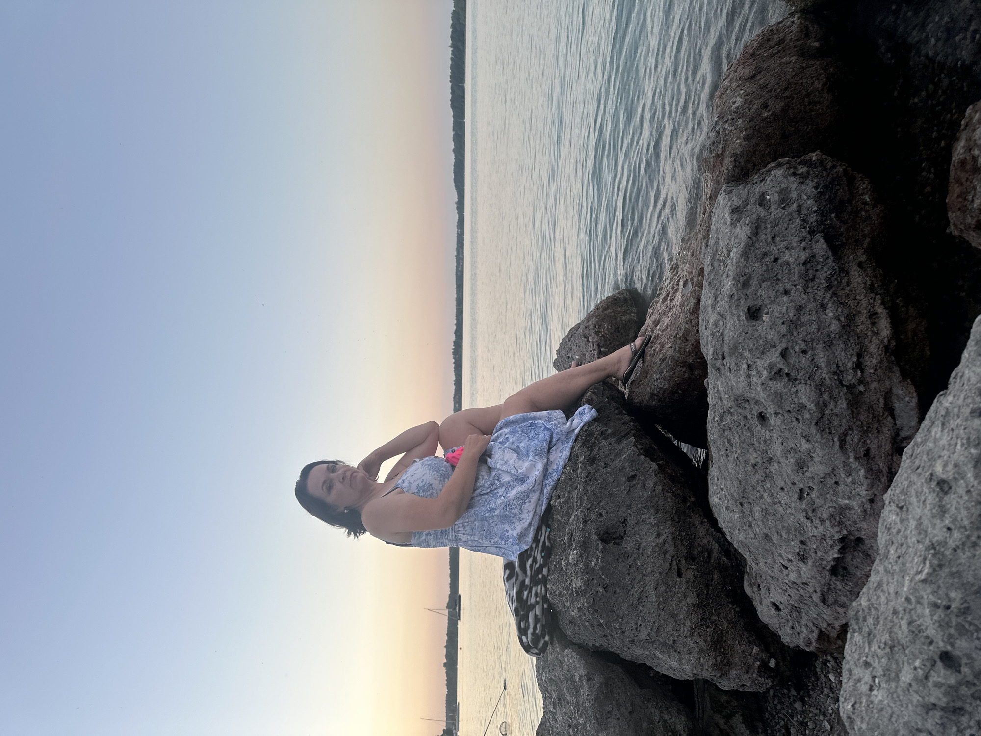A woman is sitting on rocks by the water at sunset. She is wearing a blue and white dress and has her hand on her chin. The sky is a light blue and pink, and the water is calm. There are sailboats in the background. The location is the shore of the Chiemsee in Prien am Chiemsee, Germany.