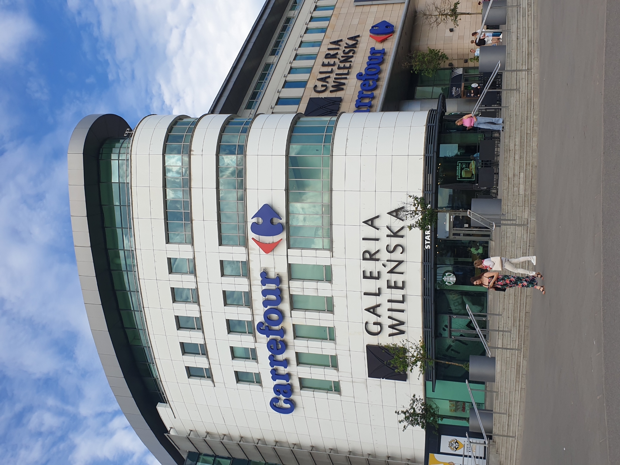 This is a vertical, eye-level, outdoor shot of the Galeria Wilenska shopping mall in Warsaw, Poland. The building has a modern, curved facade with white paneling and large windows. The "Carrefour" logo is prominently displayed in large blue letters. Above it, "GALERIA WILENSKA" is written in black letters.
Several people are visible outside the mall. Two women are standing on the steps leading up to the entrance of a Starbucks. One woman is wearing a floral dress, and the other is wearing a pink top and light-colored pants. Another woman in a pink top and jeans is standing on the steps, leaning on a railing. In the background, a few other individuals are seen walking or standing.
The sky is blue with scattered white clouds, suggesting daytime with pleasant weather. The scene appears to be set on a bright, sunny day. The building's architecture and the branding of the businesses indicate a commercial, urban environment. No specific event is discernable, and the general atmosphere seems casual and relaxed.