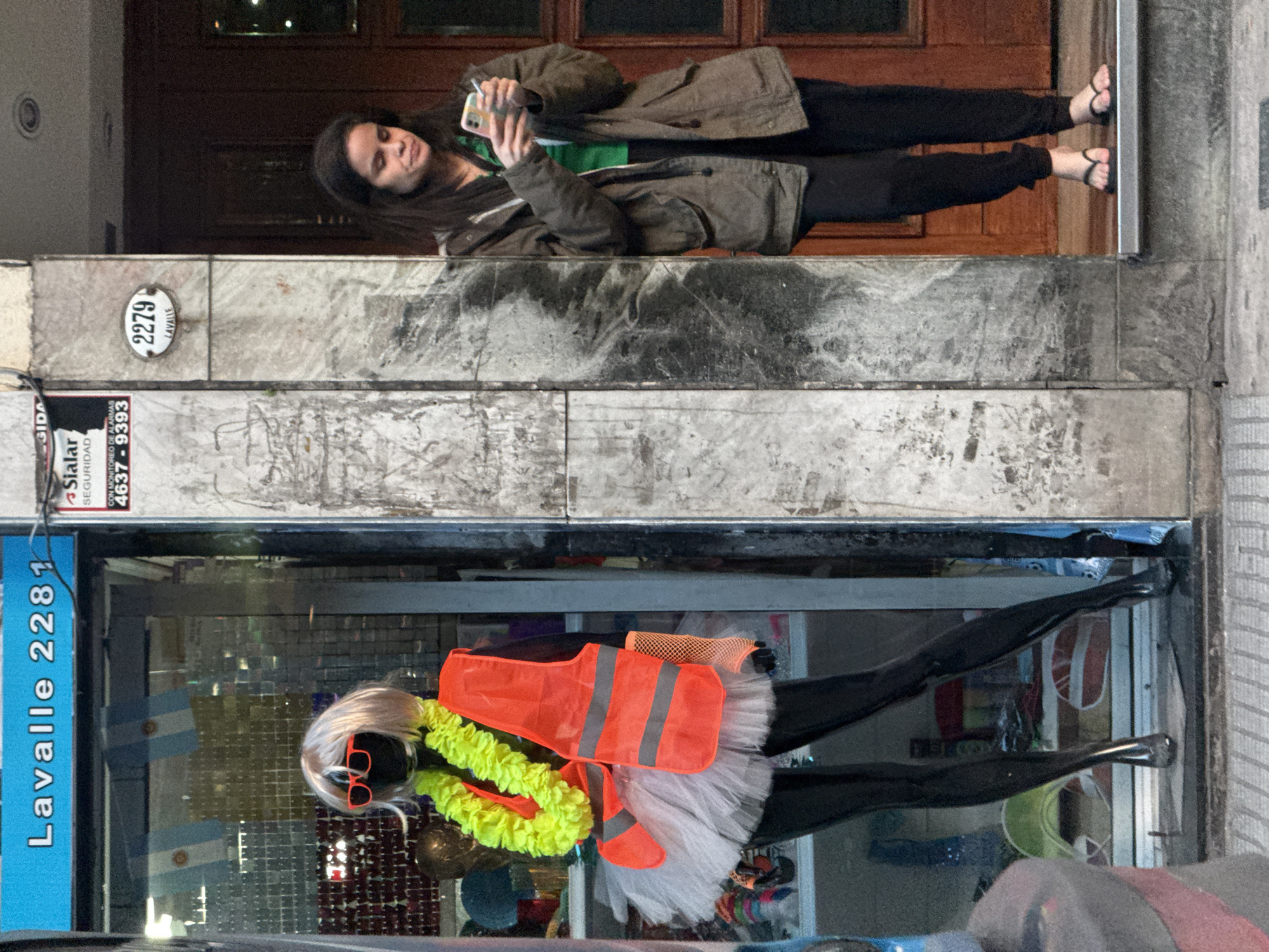 A woman stands outside a shop at Lavalle 2279/2281 in Buenos Aires, Argentina. The woman is light-skinned with long brown hair, wearing black pants, black flip-flops, a green shirt, and an olive-green jacket. She is holding and looking at a cell phone. The shop window displays a mannequin wearing black leggings, a white tutu, an orange safety vest, a yellow lei, and red sunglasses. Argentinian flags are visible in the shop window. Advertisements are pasted above the shop window.