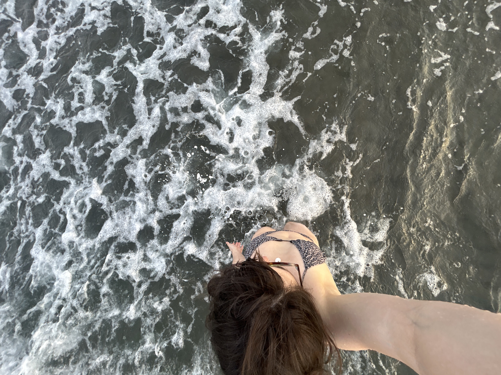 A high-angle shot captures a person's arm and back as they sit in dark, choppy water. The water is filled with white foam and waves. The person's hair is dark and long, and they are wearing a leopard-print bikini top. The scene appears to be a beach or coastal area in Ortaca, Turkey.
