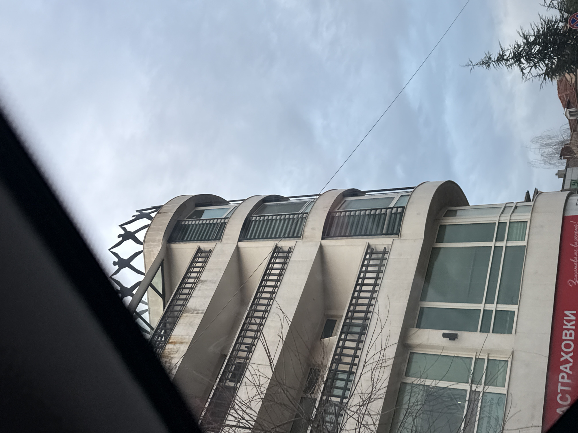 A low-angle shot captures the façade of a building in Burgas, Bulgaria. The building features a modernist architectural style with curved concrete balconies and what appear to be decorative metal ladders running vertically along the exterior. The balconies have metal railings. The building also has large windows. A sign with the word "СТРАХОВКИ" (insurance) is visible. At the top of the building, bird-like decorations are present. Bare tree branches obscure some of the building. The sky is overcast. The photograph seems to have been taken from inside a vehicle.
