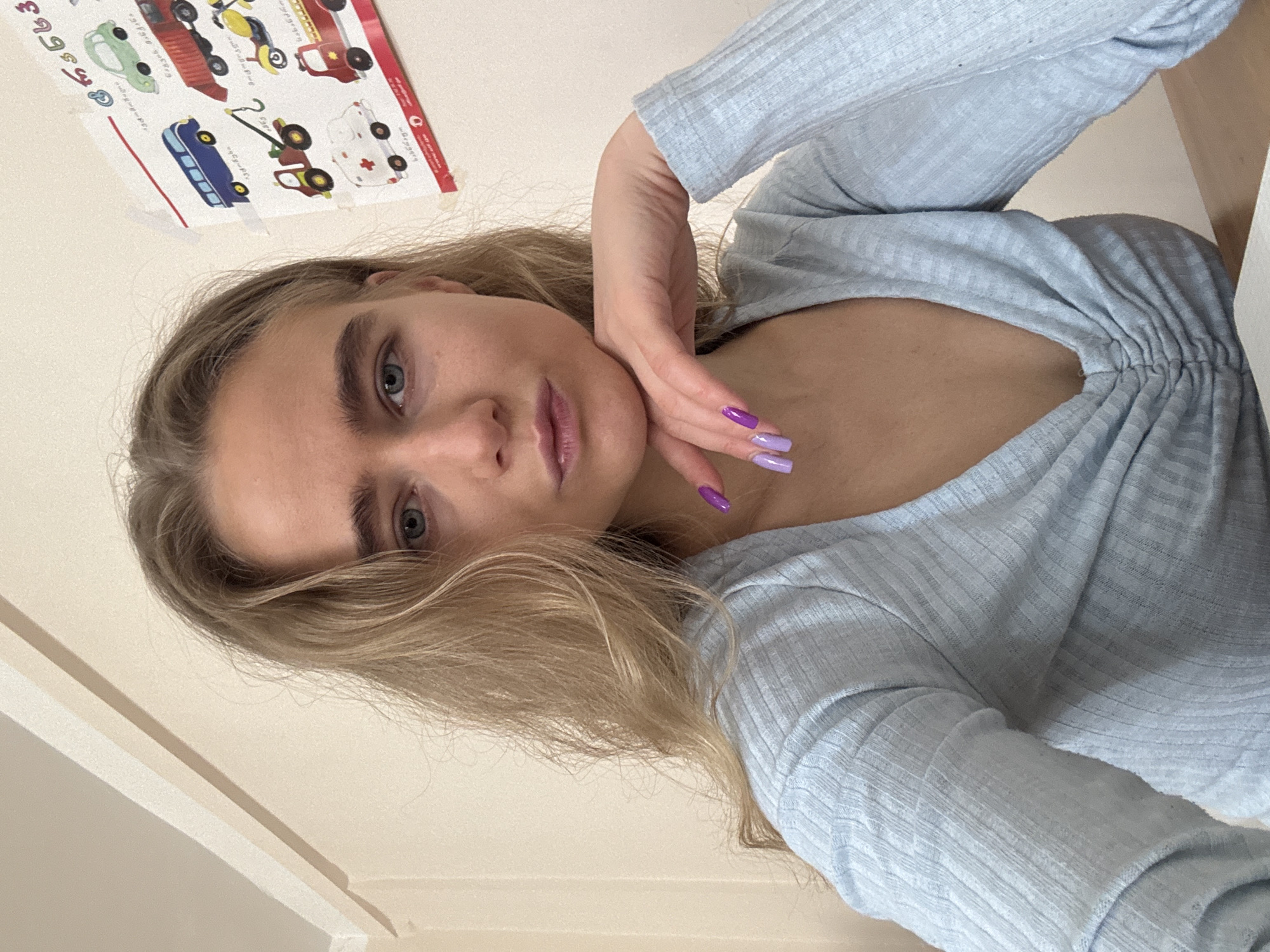 A young woman with blonde hair and light blue eyes is positioned in the center of the frame, leaning her head on her right hand with purple nail polish. She wears a light blue, fitted, long-sleeved top that is gathered at the chest. The angle of the photograph is from slightly below her, looking up. In the upper left corner of the image, a poster depicting various vehicles, such as a bus, fire engine, ambulance, and other types of vehicles, is affixed to the wall. The background is a light, creamy color, with the suggestion of a wall and ceiling meeting in the lower left corner. The location and city are unknown.