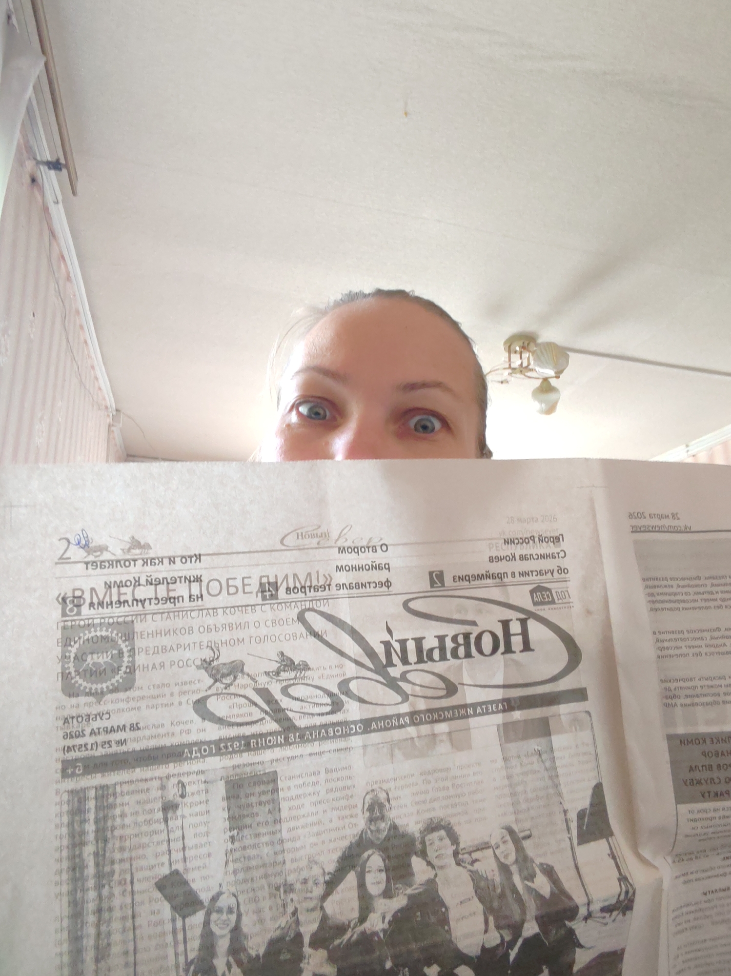 A woman is holding a newspaper with the headline "HOBOSTI" (News in Russian) in front of her face, so that only her eyes are visible. The newspaper features articles and images, including a group photo of people. The text includes "HEROI ROSSII STANISLAV KOCHEV S KOMANDOI" (Hero of Russia Stanislav Kochev with his team), indicating a political context related to elections or participation in the "EDINAYA ROSSIYA" (United Russia) party. The date "28 marta 2026" (March 28, 2026) is visible on the newspaper.
The setting appears to be indoors. The walls have a light-colored patterned wallpaper. A ceiling light fixture is visible in the background. Based on the OCR text "LVBELV NEWCKOLO DUNOHV OCHOBVHW TO NOHY TONS LOTV Radungan Diι των 20", the location is inferred as Чухлэм, Russia.