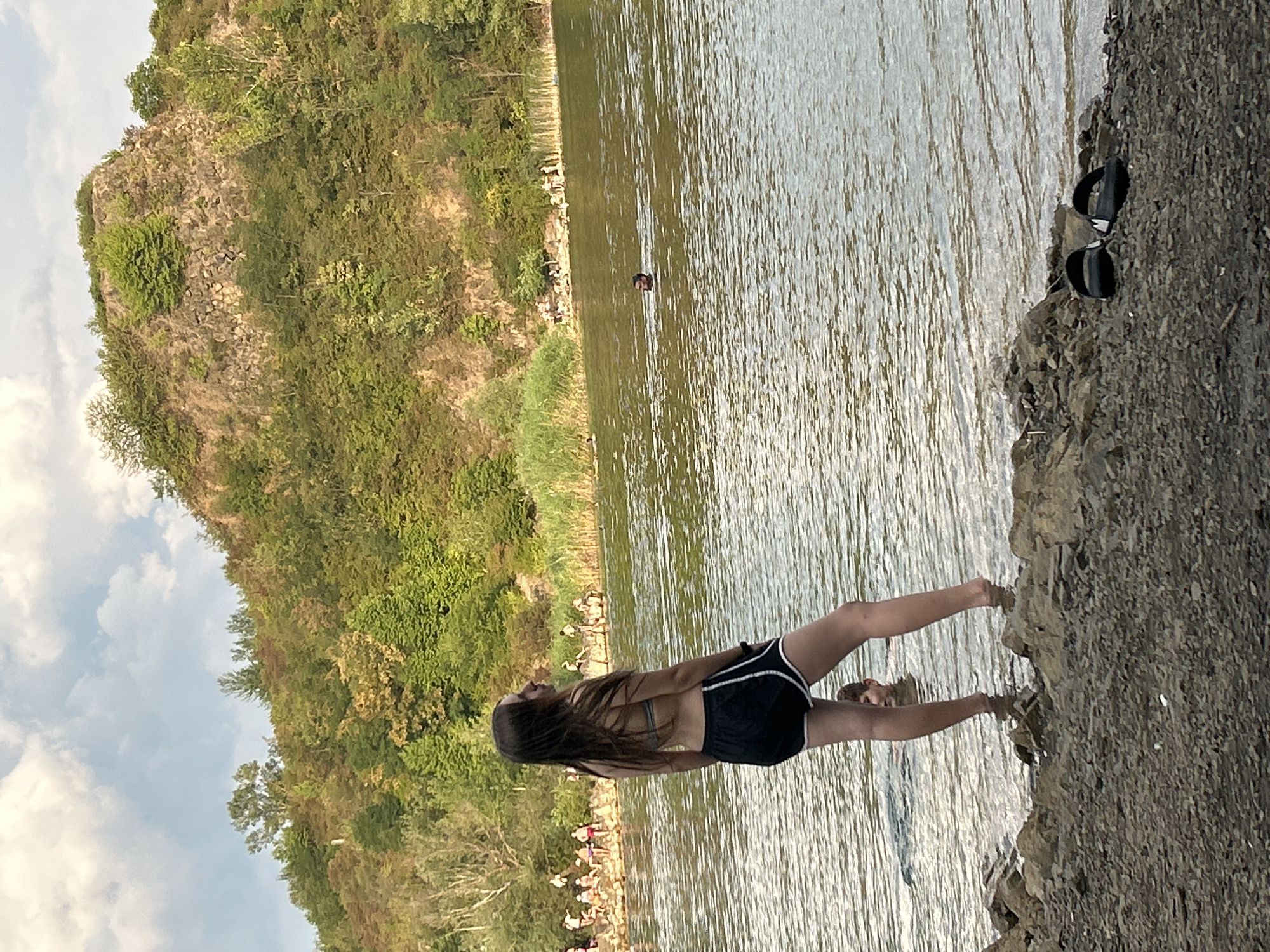 In Uzhhorod, Ukraine, on a rocky shore of a body of water, a young woman with long brown hair stands facing away from the camera, looking out at the water. She is wearing a light-colored bikini top and black shorts with white trim. Her feet are in the shallow water at the edge of the shore. Behind her, the water is rippled and reflects the light. In the distance, across the water, a tree-covered hillside rises, with a rocky cliff face visible at the top. There are several people swimming and relaxing on the opposite shore. A pair of black slide sandals rests on the rocky ground in the foreground.