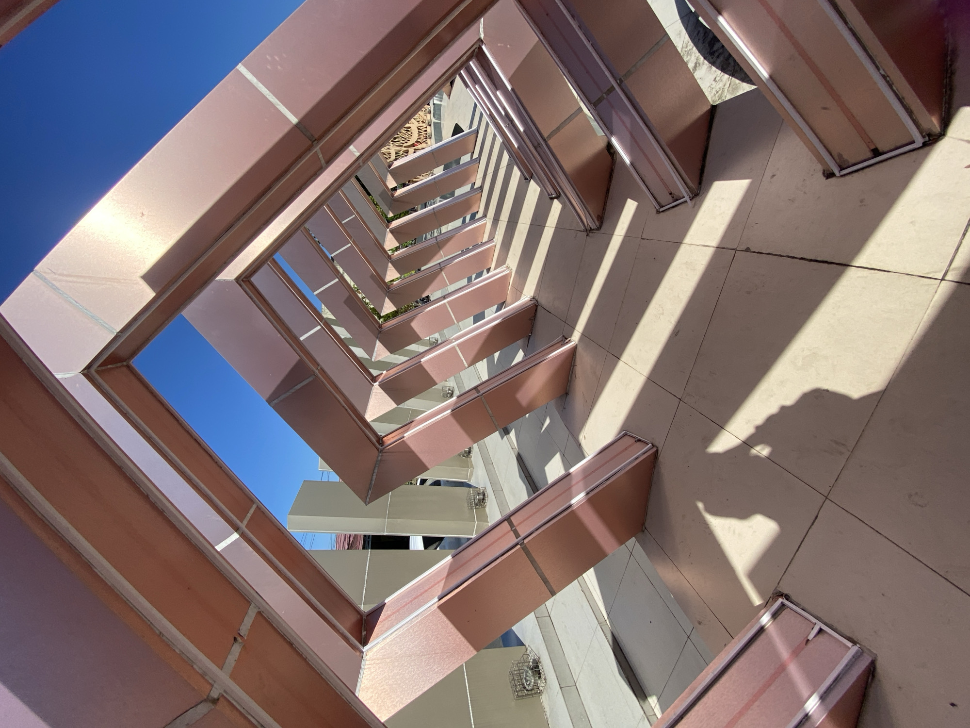 In the city of Banjarbaru, Indonesia, an outdoor perspective shot captures a modern metal sculpture on a sunny day. The sculpture, made of rose gold colored panels with white trim, forms a series of interconnected diamond shapes that recede into the distance, creating a tunnel-like effect. Sunlight casts sharp, geometric shadows from the sculpture onto the tiled ground. Beyond the sculpture, a modern building with beige and grey facades is visible, along with a clear blue sky. The overall scene is one of urban contemporary design.
