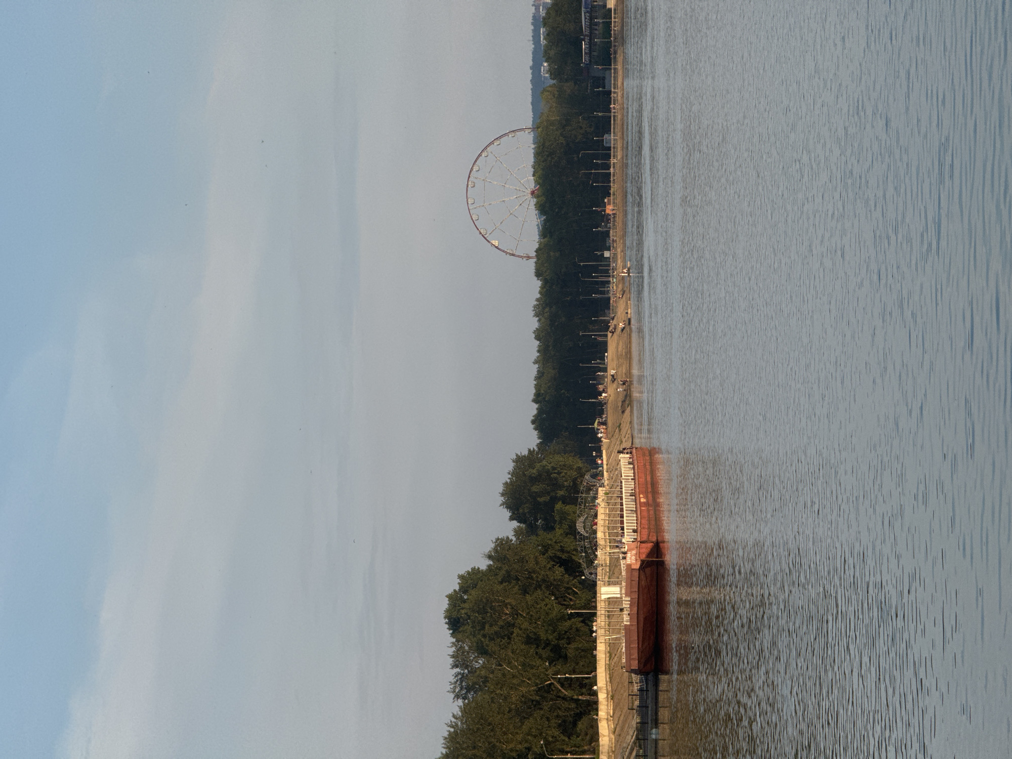 The image depicts a wide, calm body of water, likely a river, in the foreground. On the far bank, a tree-lined embankment is visible, with a sandy beach area where several people are casually gathered. Beyond the trees, a large Ferris wheel is partially visible, a prominent landmark. In the left portion of the image, a long, red barge or boat is moored to the embankment. The embankment itself is reinforced and lined with what appears to be a walkway or promenade. The sky above is a pale blue with wispy clouds. The overall scene suggests a public recreational area within a city, likely during the daytime, given the lighting. This scene is consistent with the waterfront of Irkutsk, Russia, particularly areas along the Angara River where such promenades and recreational facilities, including a Ferris wheel, are common.