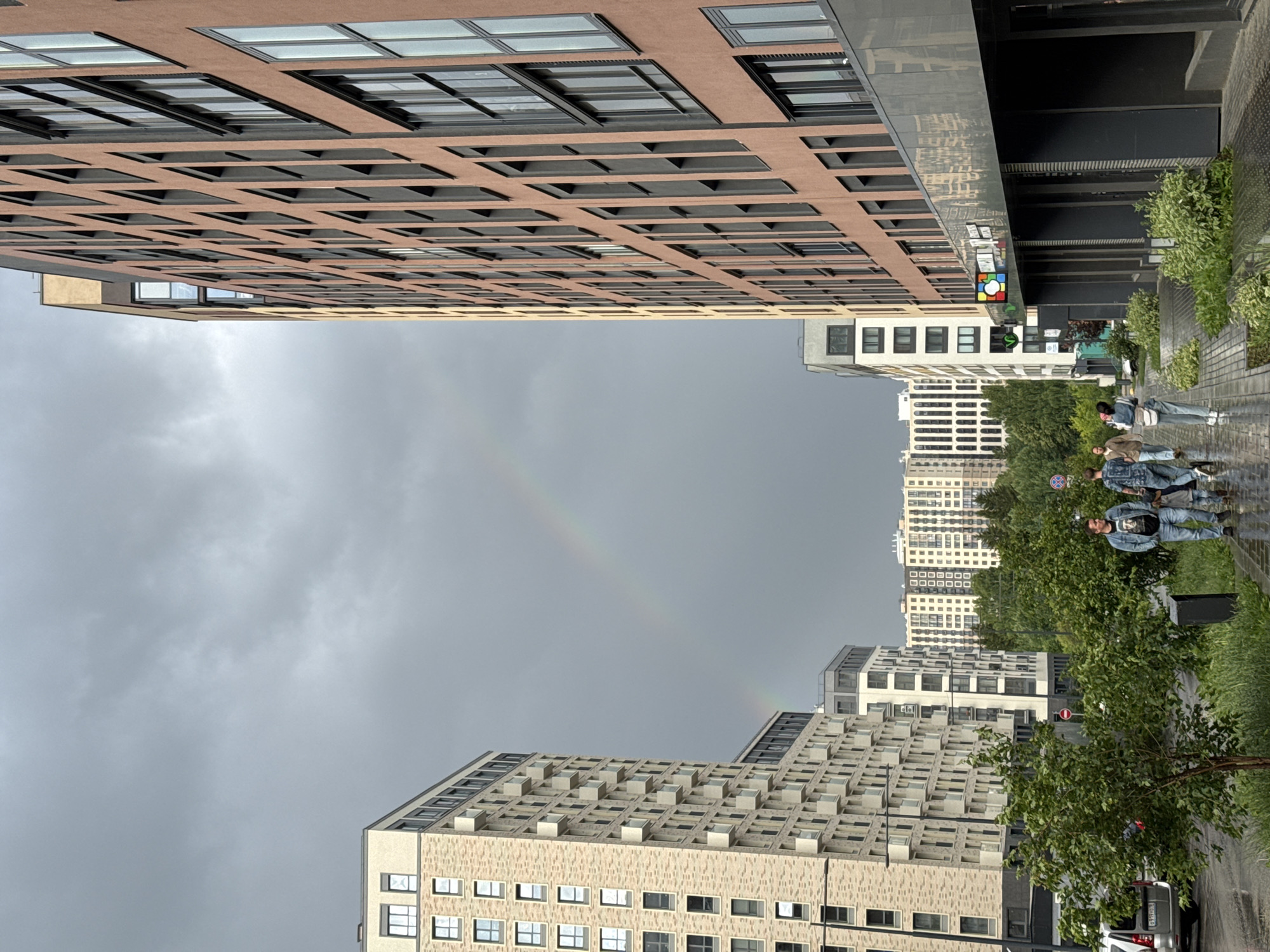 The image depicts a street scene in Izhevsk, Russia, with modern residential buildings on both sides of the street. The sky is overcast with a visible rainbow. On the left, a tall, light-colored building with many balconies dominates the foreground. On the right, a taller, reddish-brown building with a grid of windows runs along the street. Several people are walking on the sidewalk in the middle ground. The sidewalk appears wet, suggesting recent rain. Trees and greenery are visible between the buildings. In the background, more residential buildings and trees are present. A white car is parked on the left side of the street.