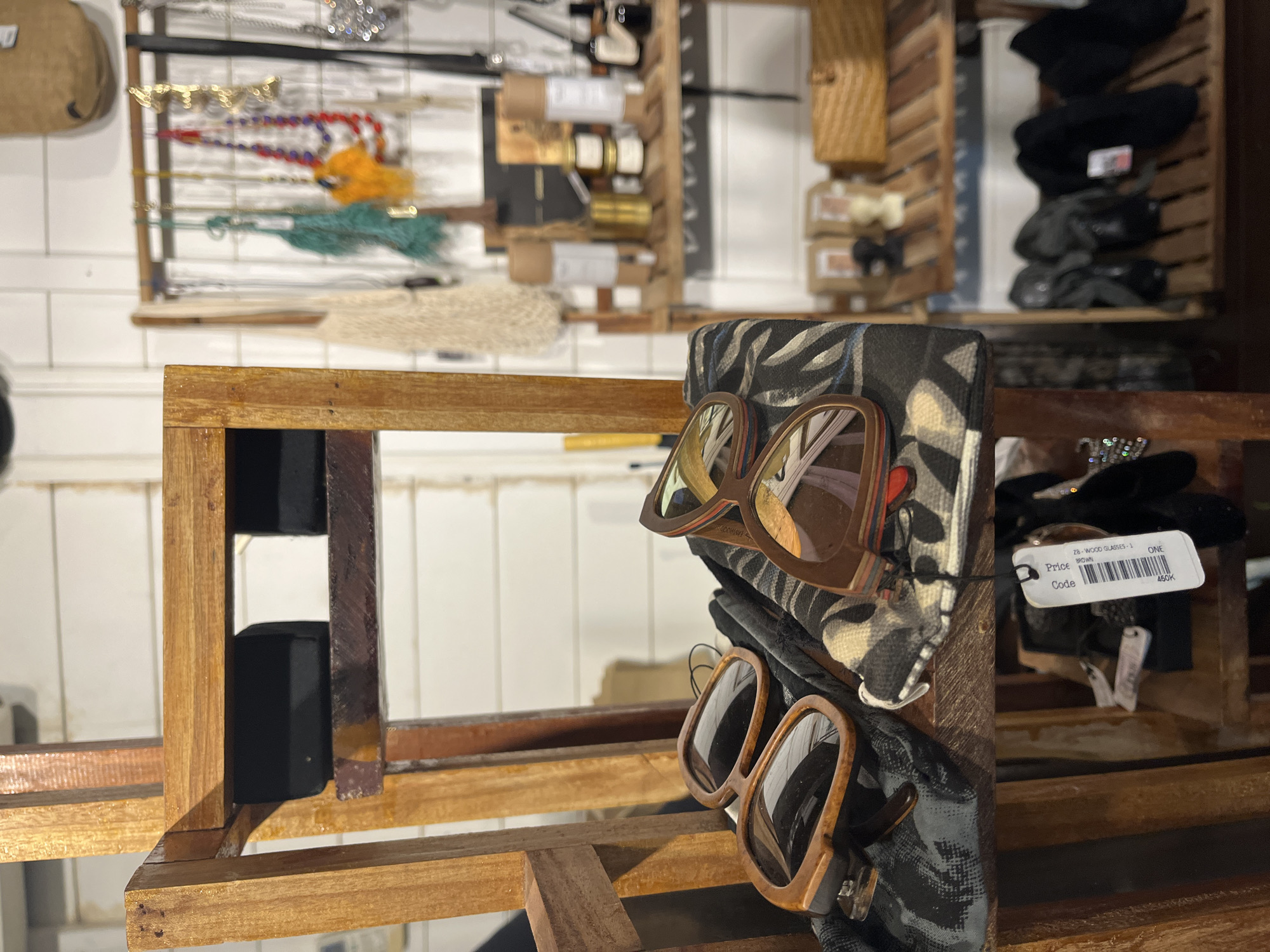 This is a medium shot, eye-level, indoor photograph of a retail display, likely in a shop in North Kuta, Indonesia. The focal point is a wooden shelf unit holding various items. In the foreground, resting on a patterned fabric, are two pairs of sunglasses with brown frames. A price tag for "28-WOOD GLASSES 1 BROWN" is visible. Behind the sunglasses, on higher shelves of the same wooden unit, are black objects, possibly storage containers.
The background reveals additional shelves displaying an assortment of products. On one shelf, there are several small jars and bottles, along with what appear to be small decorative figures. On a higher shelf, a collection of necklaces with colorful beads and other decorative elements are hanging from a rack. Further to the right, a woven basket is visible. The lowest shelf shows several pairs of dark-colored footwear. The walls appear to be white and paneled. The lighting is natural and somewhat diffused, suggesting an indoor retail environment. There are no people visible in the image.