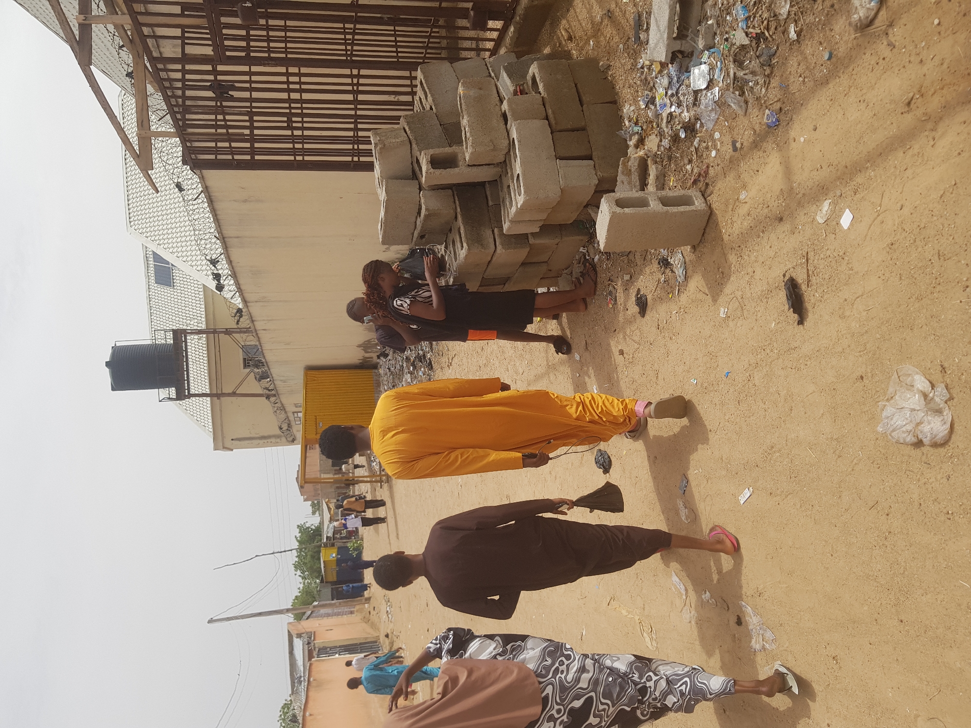 The image shows a street scene in what appears to be Dabawa, Nigeria. Four people are visible. Two individuals stand by a stack of concrete blocks next to a building with a fenced rooftop. One of them is holding what appears to be a phone. Another person is walking, wearing a yellow robe and holding a black object. Further down the street, a person in a brown garment is also walking, followed by someone wearing patterned pants. The ground is sandy with scattered debris. Buildings are visible along the street in the background.