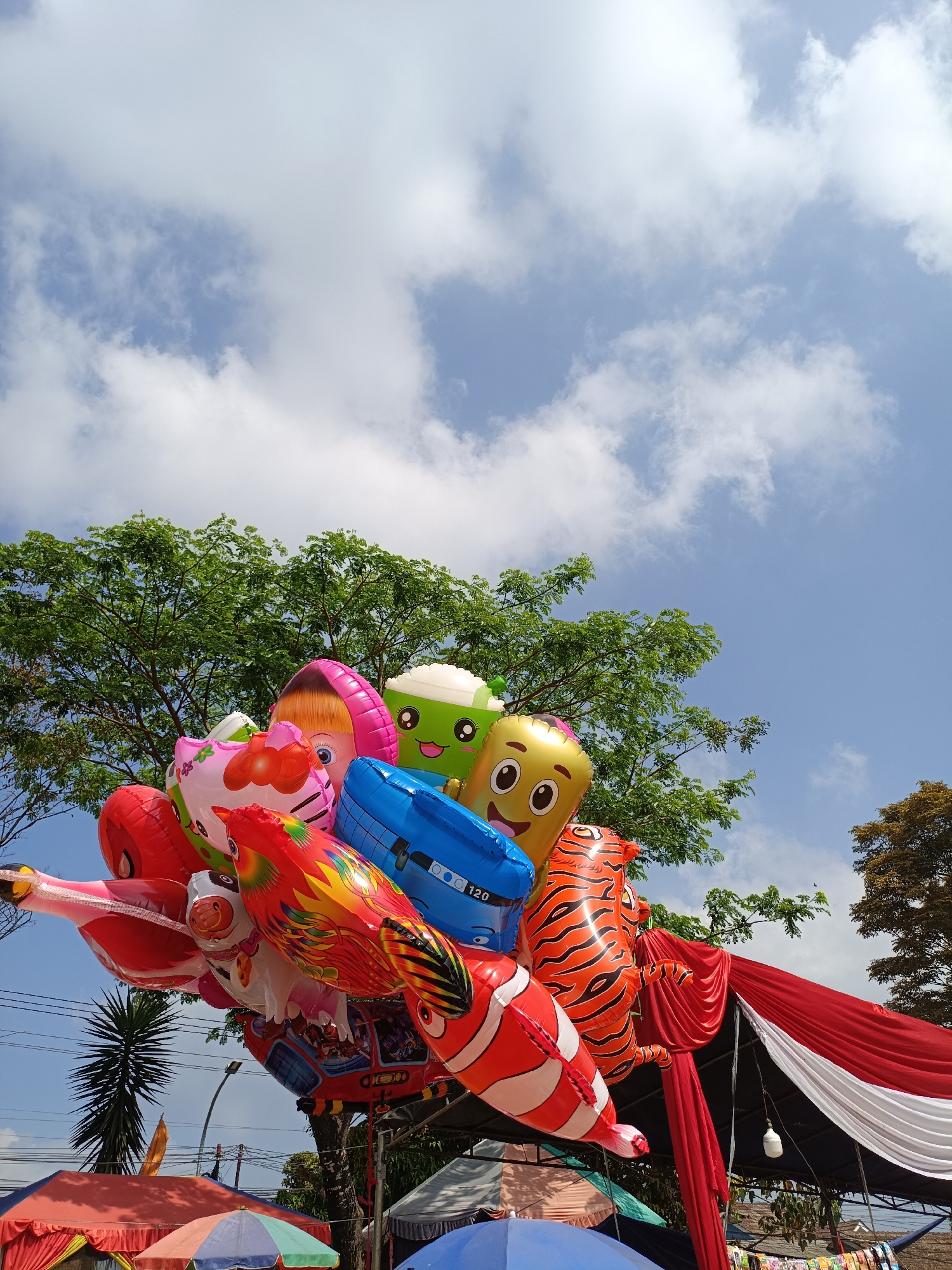 A low-angle shot captures a cluster of colorful inflatable balloons against a bright blue sky with scattered white clouds, framed by lush green trees and foliage. The balloons are diverse in shape and design, featuring cartoon characters, animals like a clownfish and a tiger, and a chicken. Some balloons are red, pink, yellow, green, blue, and orange. A red and white draped fabric, possibly a banner or decoration, hangs in the background, along with electrical wires and poles. Below the balloons, glimpses of market stalls with red and blue awnings are visible, suggesting an outdoor event or marketplace. The scene appears to be in an urban or semi-urban outdoor setting, with a clear, sunny day. The location is identified as Cibodas, Indonesia.