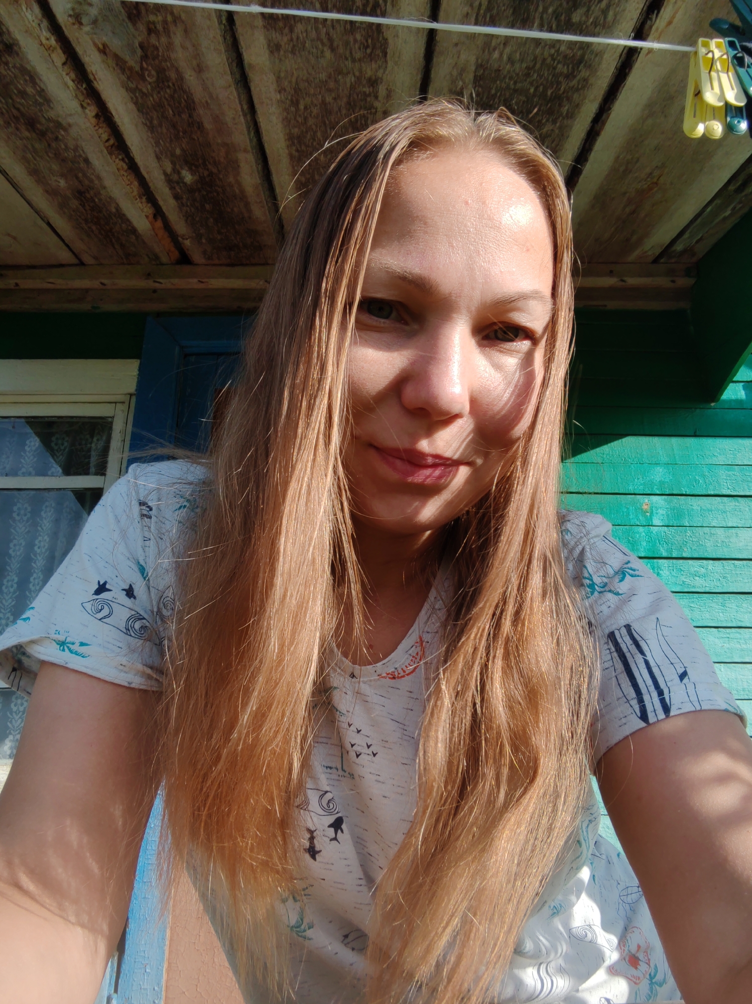 A young woman with long, blonde hair is taking a selfie in front of a teal wooden wall and a wooden porch ceiling. She is wearing a light-colored t-shirt with a pattern of marine life and birds. The lighting suggests it is daytime, and the environment appears to be a rural or outdoor setting, possibly in a dacha (country house). The background includes a window with blue shutters. There are yellow and green clothespins hanging on a white line above her head. The overall scene conveys a casual and relaxed atmosphere.