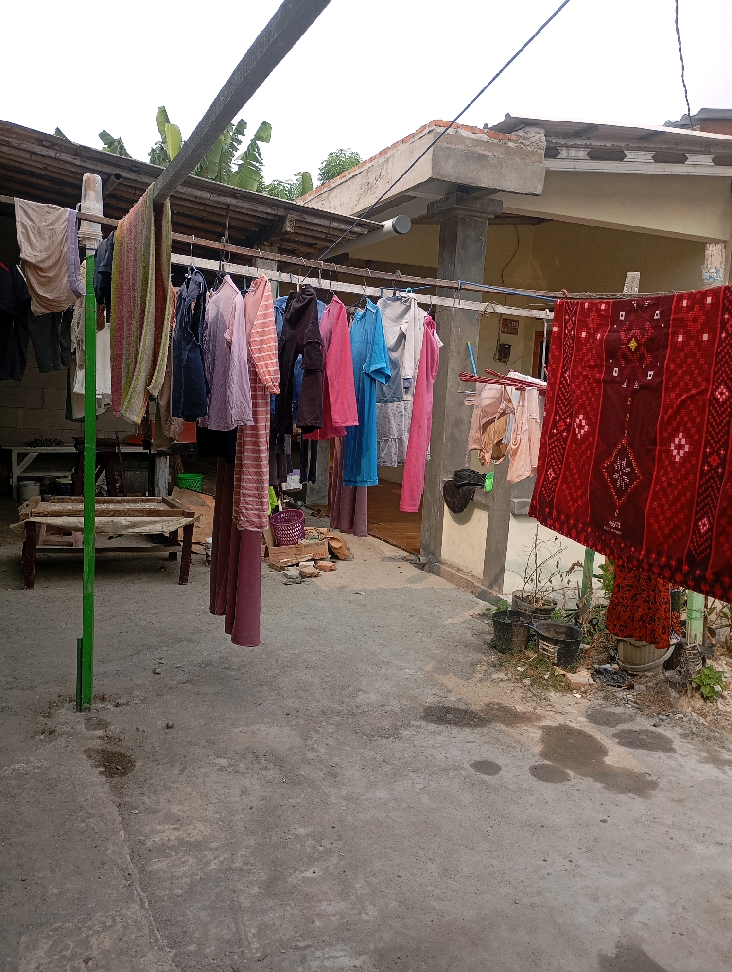 This image depicts an outdoor drying area for laundry in Babelan, Indonesia. A clothesline is strung across the frame, holding various garments including shirts, trousers, and a patterned red cloth. The laundry is hung on metal hangers. In the background, a building with a light-colored facade and a corrugated metal roof is visible. The ground is concrete, with some wet patches suggesting recent rain or washing. Several potted plants and buckets are present near the building. A wooden bench with a cloth draped over it is positioned to the left of the frame. The overall scene is one of domestic activity in a humid, tropical environment.
