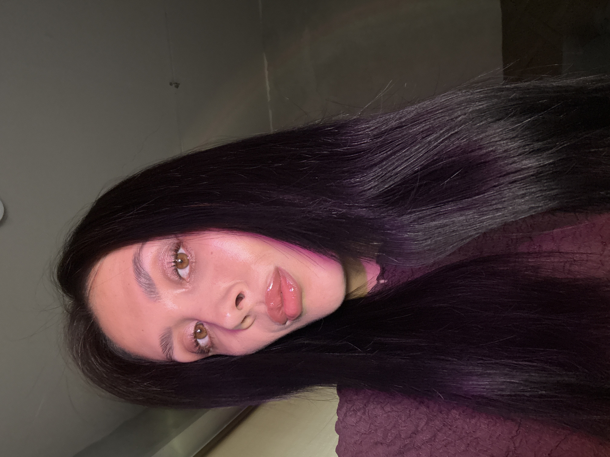 The image depicts a woman with long, dark hair, fair skin, and brown eyes. She is positioned in a way that her face is the primary focus of the shot, with her head tilted to the side. Her lips appear to be glossy. The background consists of a grey wall and a patterned purple fabric. There is no discernible indication of location in the city of Votkinsk, Russia.