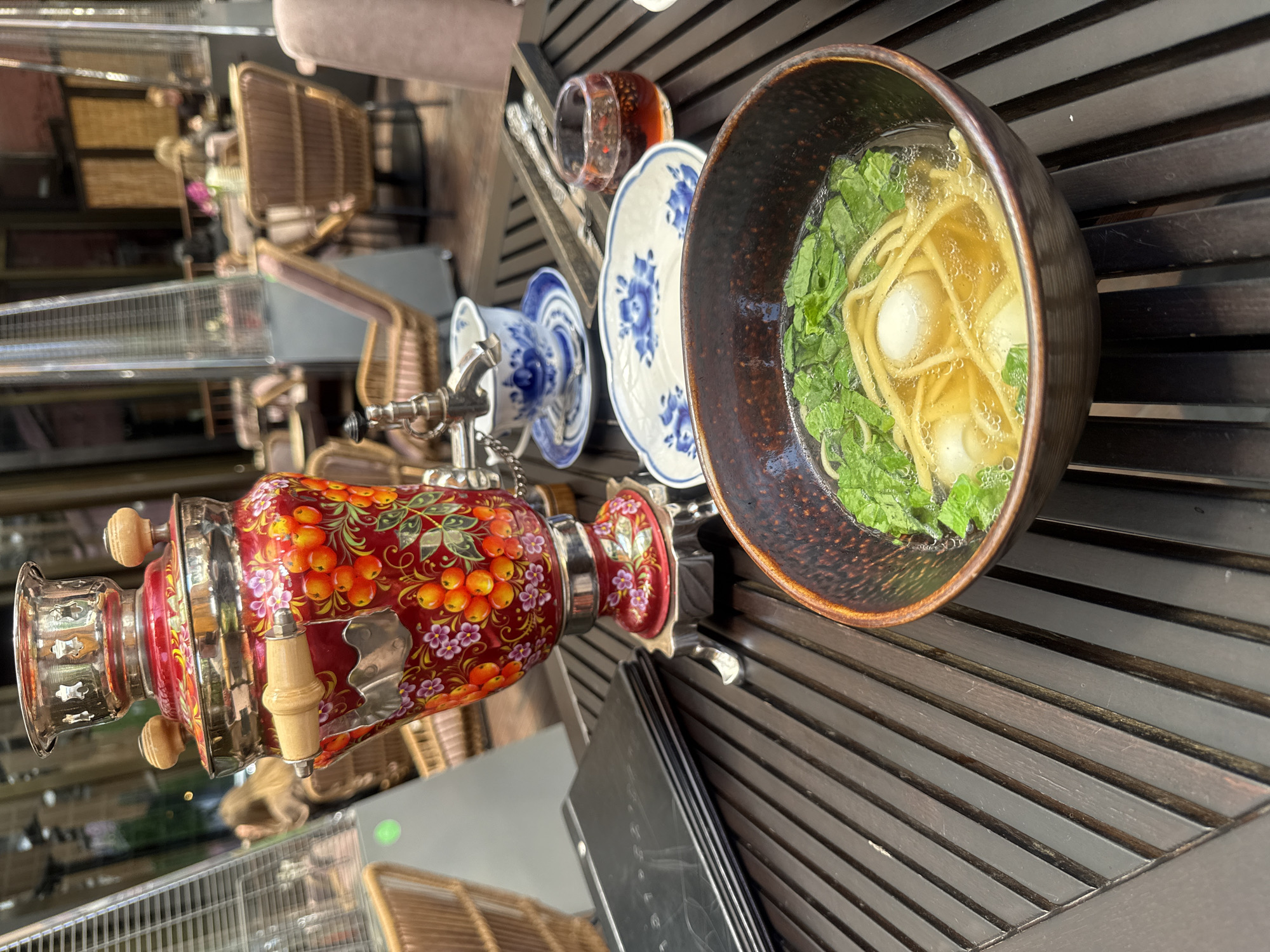 An outdoor restaurant table in Vladivostok, Russia, displays a meal. A red and gold samovar with floral and fruit motifs sits prominently on the table, its spout open. In front of the samovar is a dark bowl filled with a light broth, noodles, and green leafy vegetables, possibly garnished with quail eggs. To the right of the samovar, a delicate blue and white teacup and saucer are visible, along with a small glass of dark liquid, possibly tea or a digestif, and a set of cutlery in a wooden holder. A stack of black menus rests to the left of the samovar. In the background, wicker chairs and tall outdoor heaters suggest a pleasant, albeit cool, dining environment. Several indistinct figures are seated at tables in the background, their faces blurred. The table itself is made of dark wood planks.