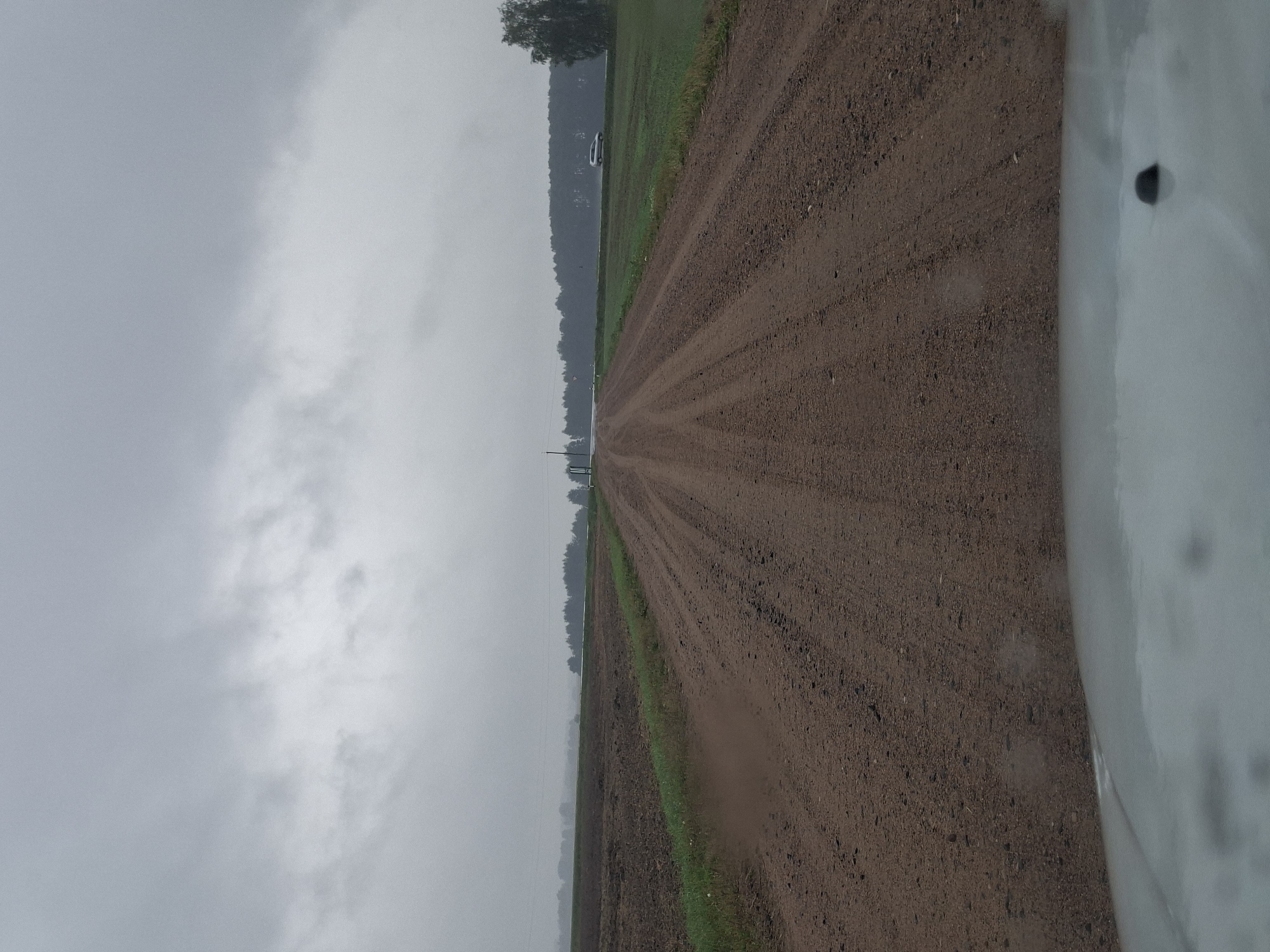 The image depicts a straight, unpaved dirt road stretching into the distance under a heavily overcast, gray sky. The road is flanked by cultivated fields and green grassy areas. In the distance, a line of trees forms a dark horizon. A white vehicle is visible on a paved road to the right of the dirt road, amidst the trees. The foreground shows the hood of a vehicle, suggesting the image was taken from inside a car. The overall scene is rural and somber, likely captured during or after rain, given the damp appearance of the road and the diffused light. The location is identified as Slavensk Rural Council, Belarus.