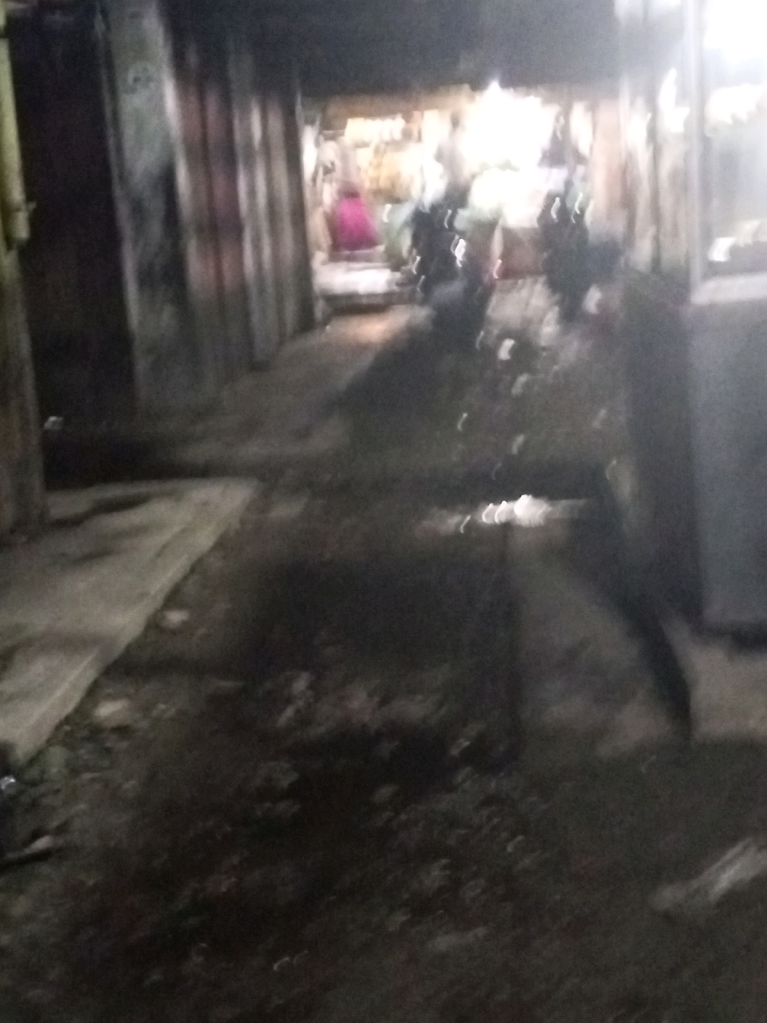 The image depicts a dimly lit, narrow alleyway or passage, possibly a market stall area, in Pancur Batu, Indonesia. The scene is characterized by wet, reflective ground, suggesting recent rain or a water source. On the left side, a textured wall, possibly corrugated metal or concrete, is visible. The right side features a brightly lit area, likely stalls or shops, with an indistinct figure in the background wearing a pink garment. Several dark, blurred shapes suggest motorcycles parked in the alley. The overall impression is one of a bustling, albeit gritty, urban environment at night.