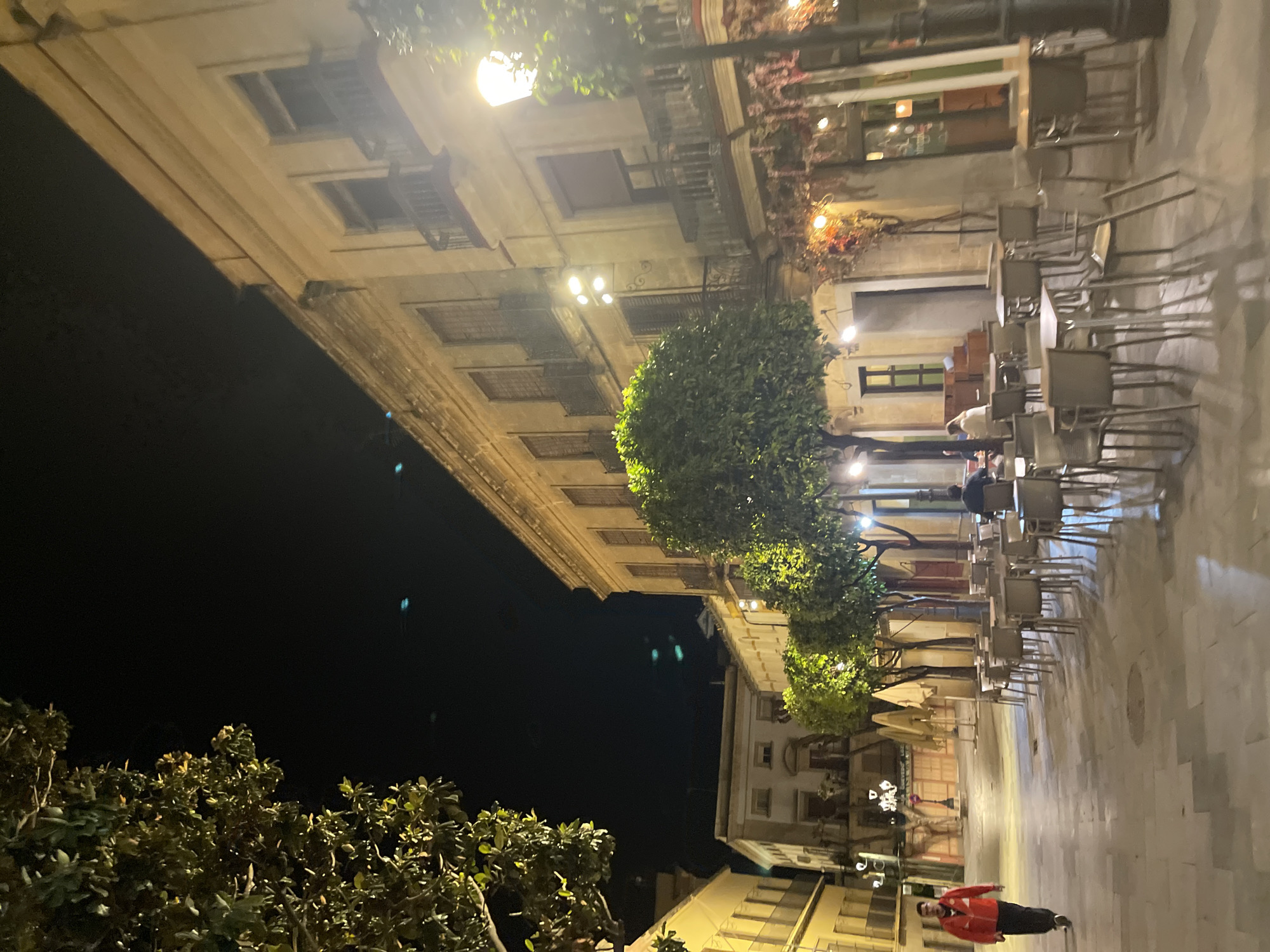 The image depicts a night scene in Jerez De La Frontera, Spain. It features outdoor seating of a restaurant with tables and chairs lined up. A few individuals are present, including a person wearing a red jacket walking towards the camera. The buildings surrounding the area are illuminated by streetlights. Trees are interspersed along the street. The scene is captured from a low-angle perspective, giving emphasis to the height of the buildings.