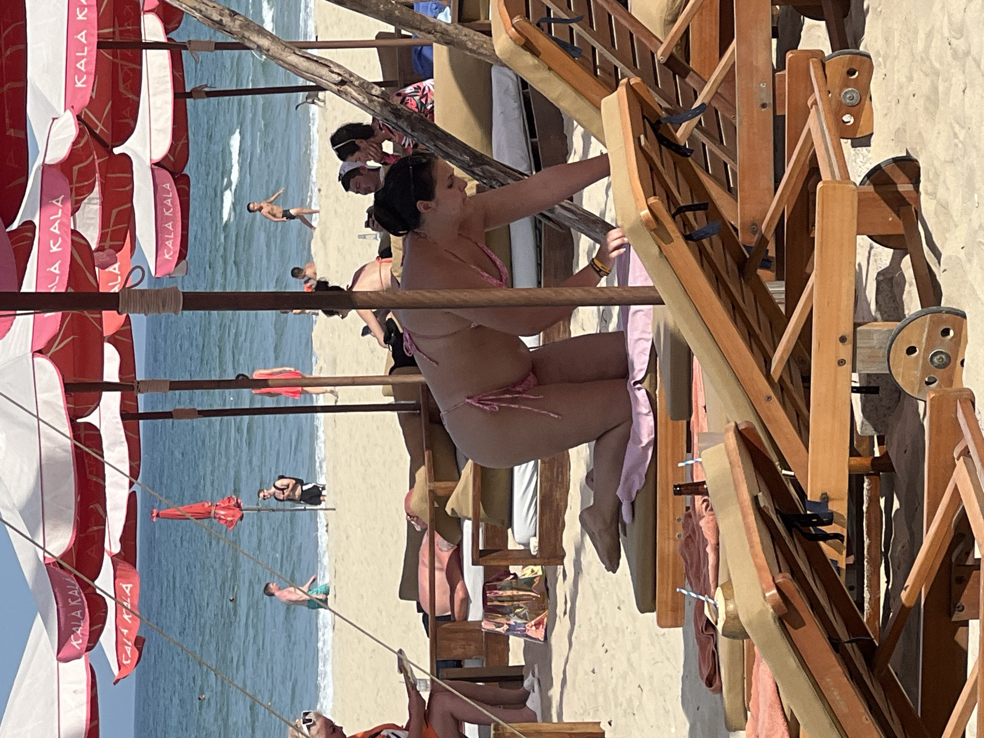 A group of people are seen relaxing on a sandy beach in front of the ocean, under large red and white umbrellas that bear the "KALA KALA" logo. The beach is lined with wooden lounge chairs, some of which are occupied. In the foreground, a woman in a pink bikini is kneeling on a lounge chair, reaching down. In the background, people are walking along the shore and swimming in the water. A few red and white beach umbrellas are set up on the sand, providing shade. The scene appears to be a tourist resort or public beach area in Đà Nẵng, Vietnam, a popular coastal city known for its beaches.