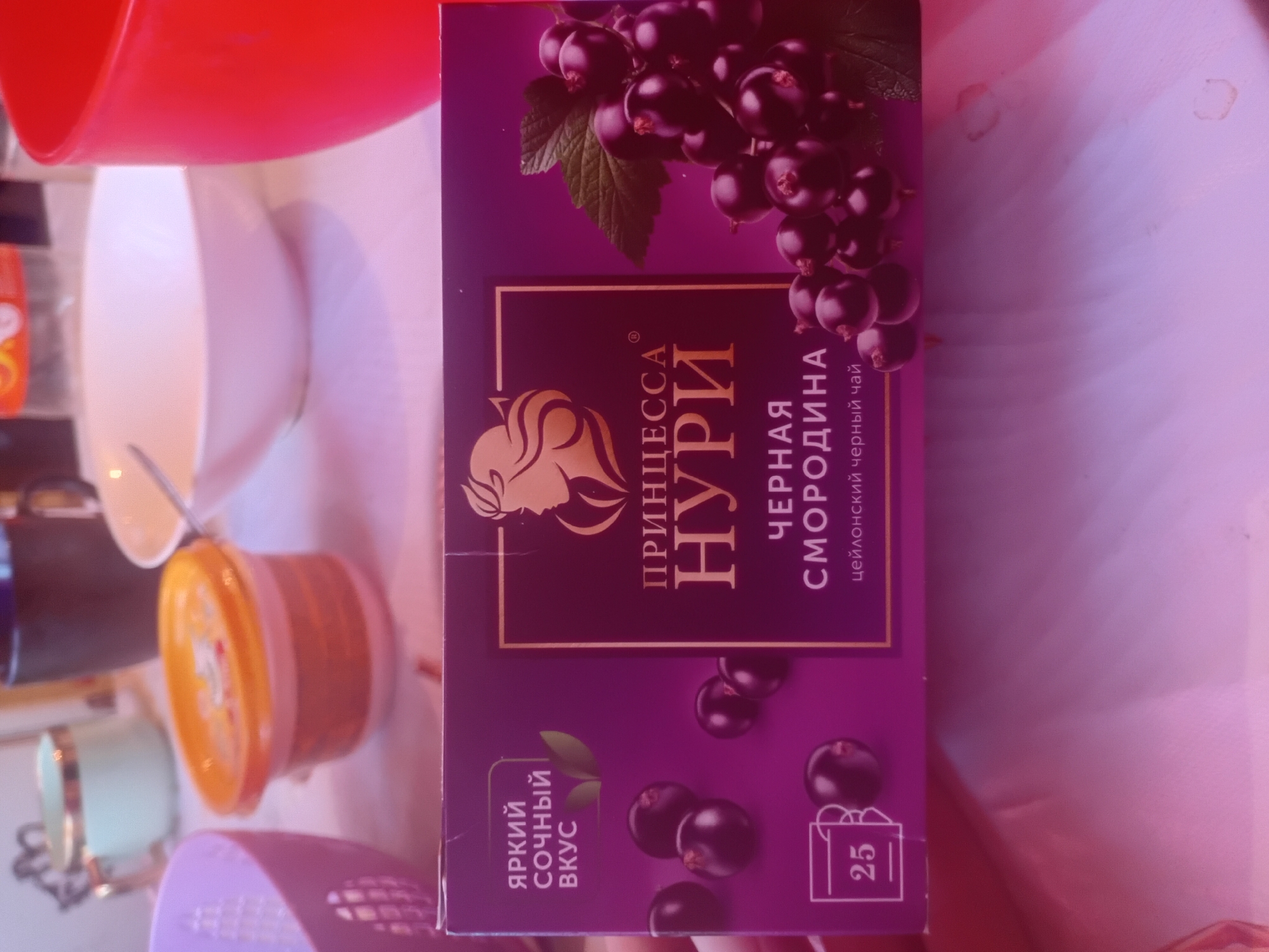 A purple tea box is pictured on a table. On the box, text in Russian reads "Princess Nuri Black Currant Ceylon Black Tea" and "Bright Juicy Taste." There are images of black currants and tea bags on the box. The box is in the foreground, and blurry objects, including cups and bowls, are in the background. The image appears to be taken indoors. The location of Малая Владимировка ауылдық округі, Kazakhstan is not evident in the image.