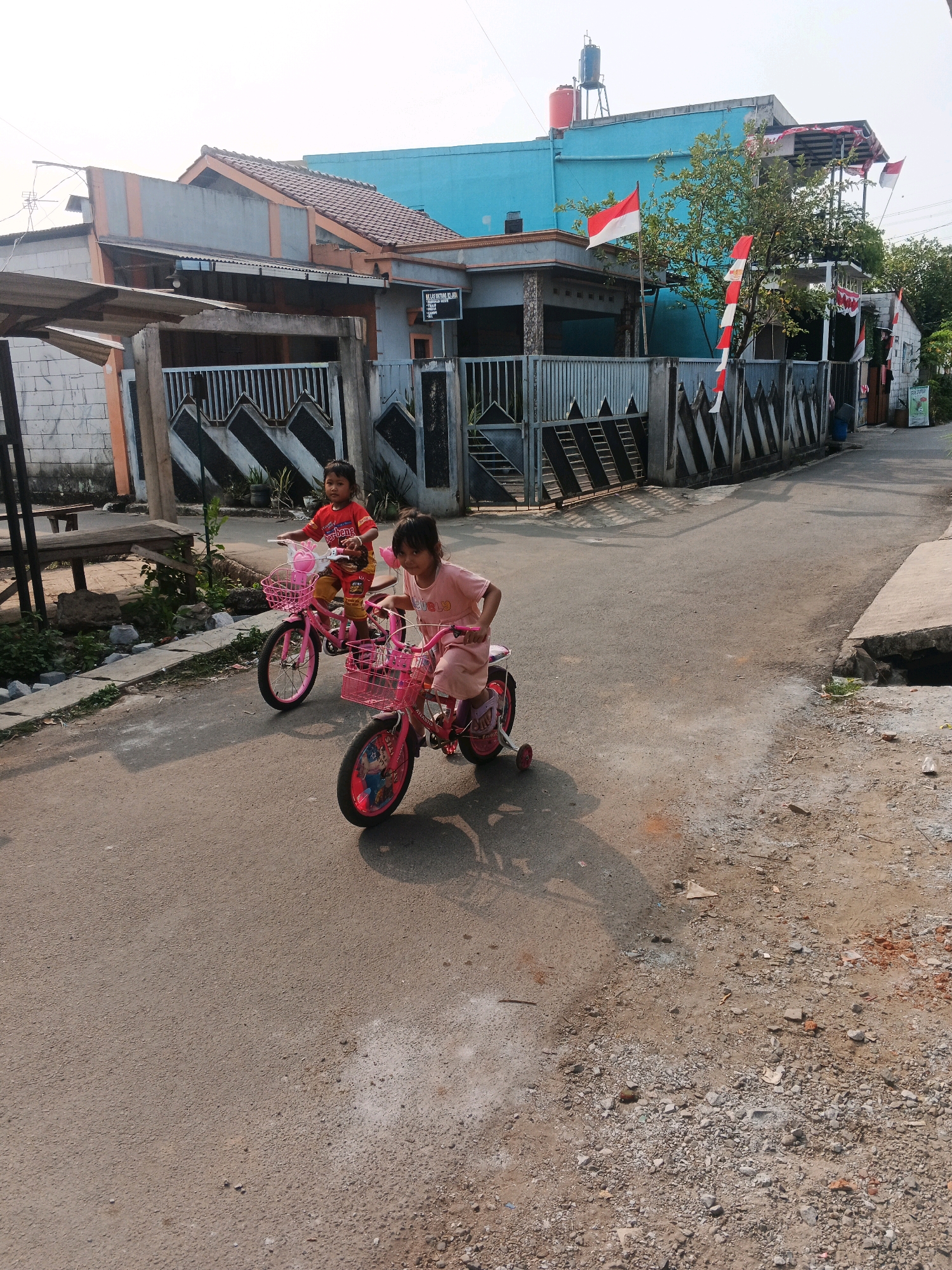 In the city of Kadu, Indonesia, two young girls are riding bicycles down a paved street. The girl on the left wears a red shirt and yellow pants, while the girl on the right is dressed in a pink dress. Both bicycles are pink and have baskets. The street is lined with houses and fences. In the background, Indonesian flags are displayed on some of the houses, suggesting a national celebration. The sky is clear and the sun is shining, casting shadows on the street.
