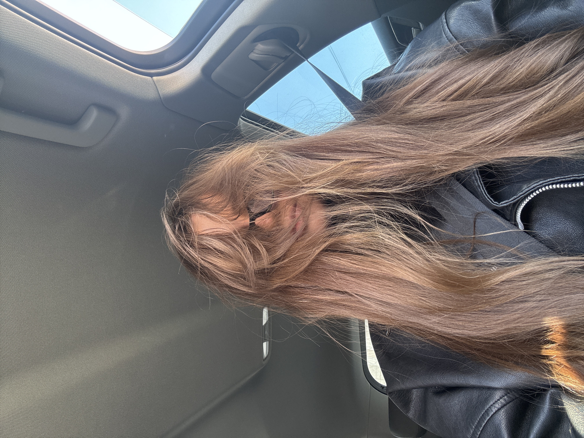 A woman with long, light-brown hair sits in a car with a sunroof. She wears glasses and a black leather jacket. The car's interior is gray. The location cannot be identified as Krasnoyarsk, Russia based on the visual information.